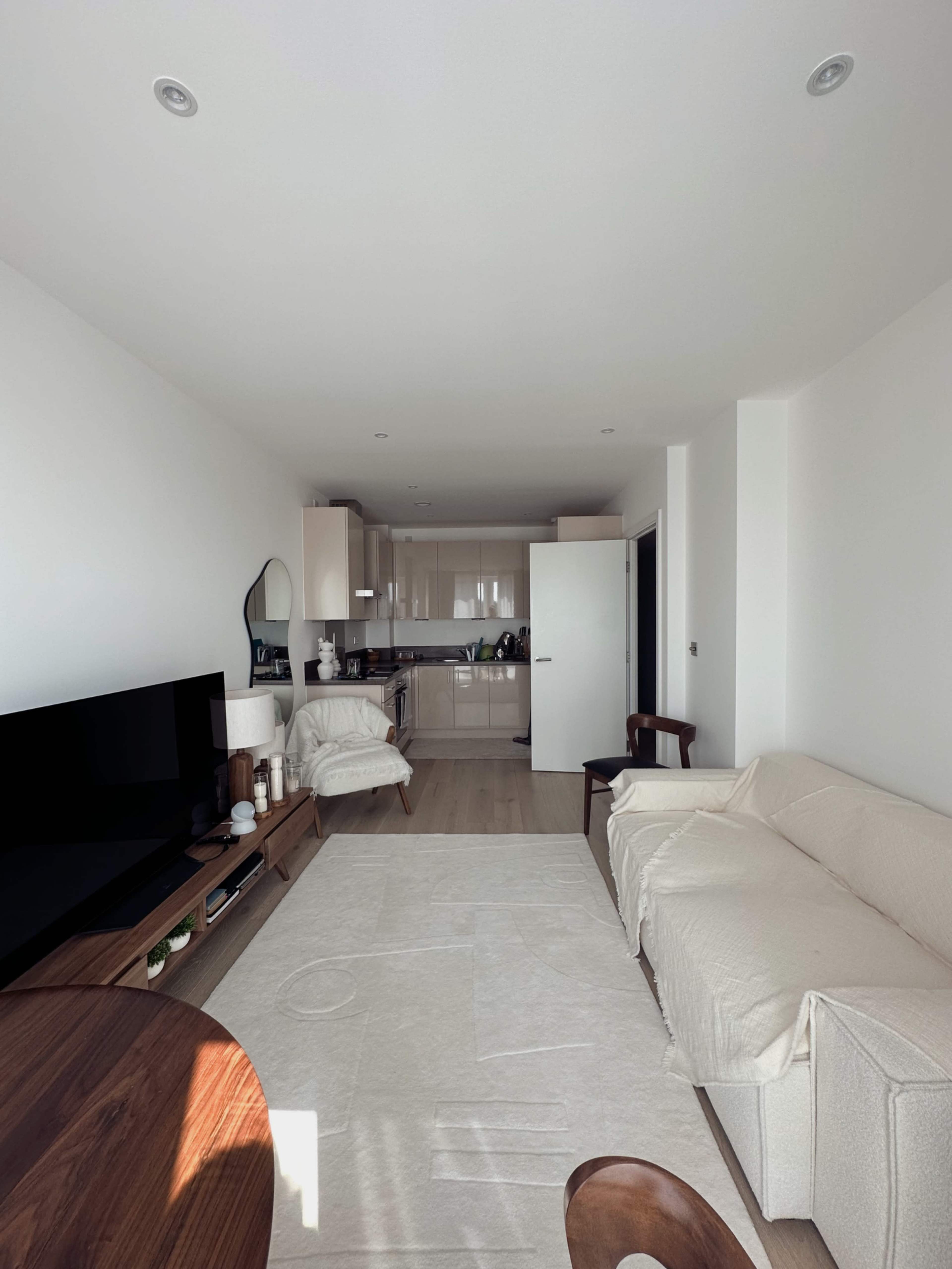 The image shows a modern, minimalistic living room with a sofa, a wooden table, a television, and an open kitchen in the background.