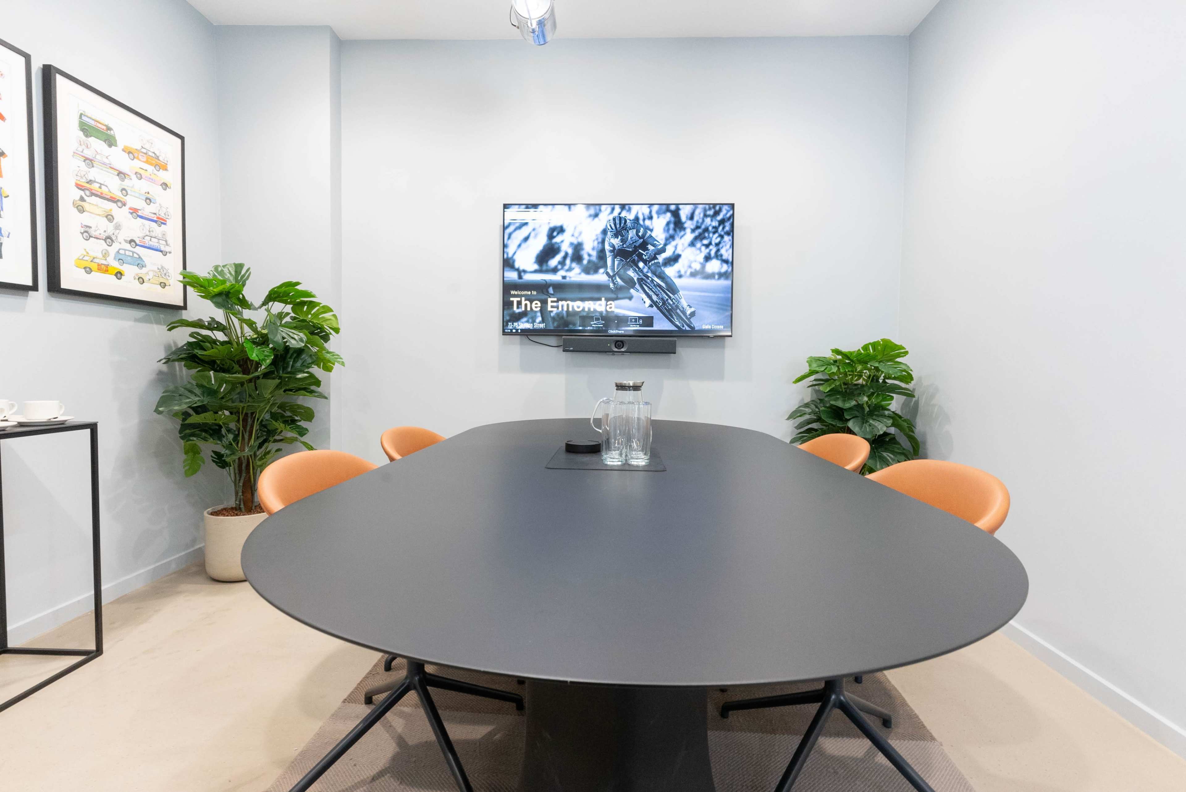 A modern conference room features a large oval table, four orange chairs, a wall-mounted TV displaying a black and white image, and two potted plants.