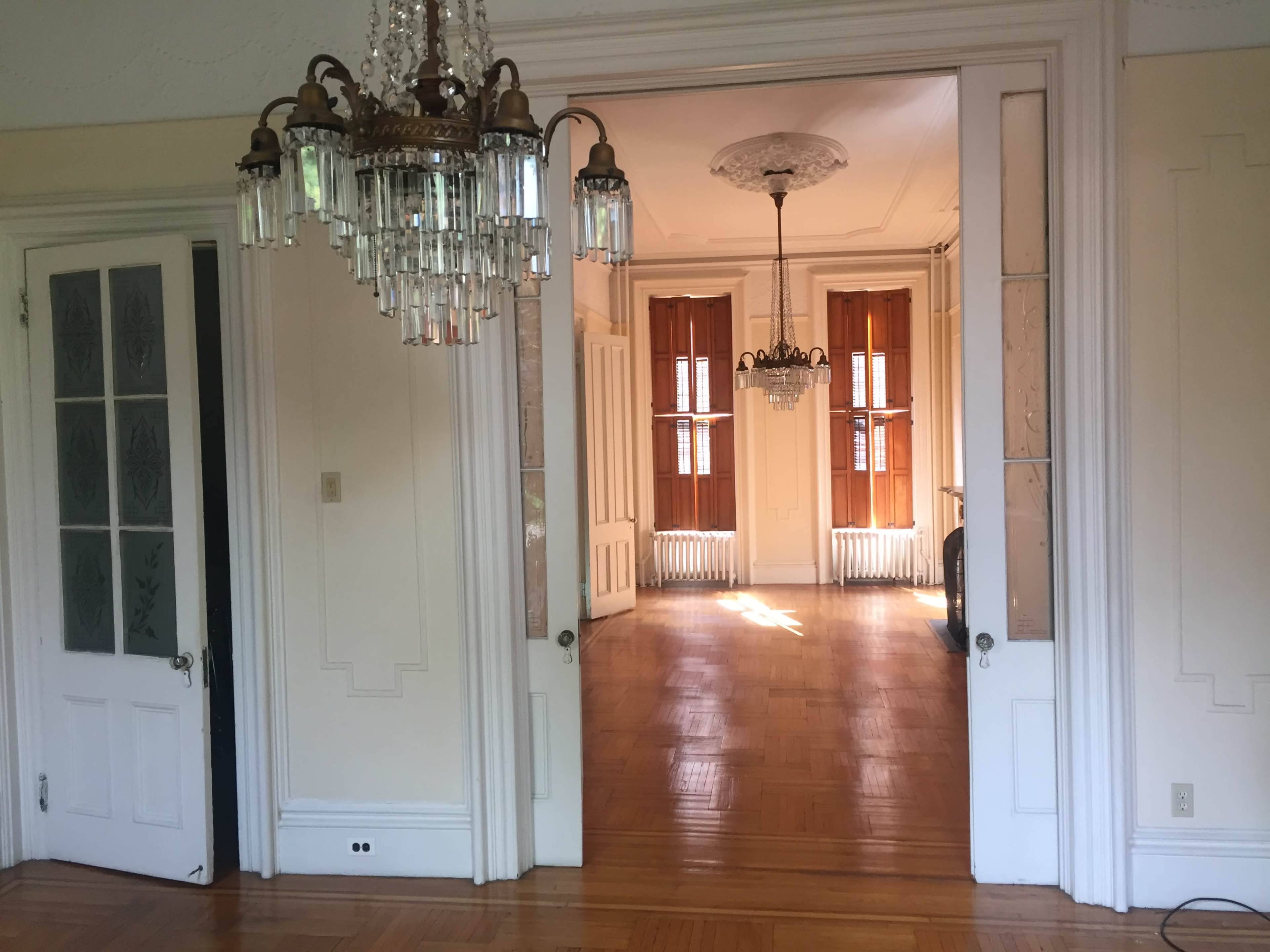 A well-lit room with a chandelier and another room visible through an open doorway, featuring large windows and hardwood floors.