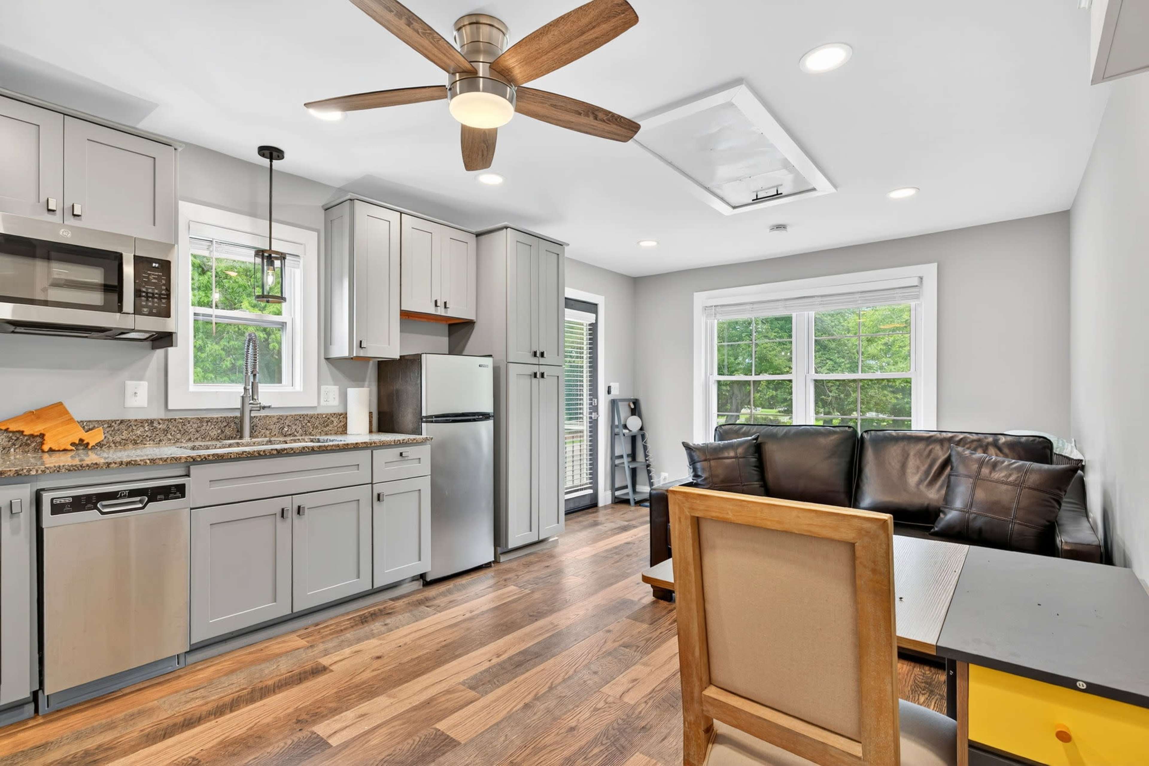 The image shows a modern kitchen area with gray cabinetry, stainless steel appliances, and a seating area with a brown leather couch and a small table.