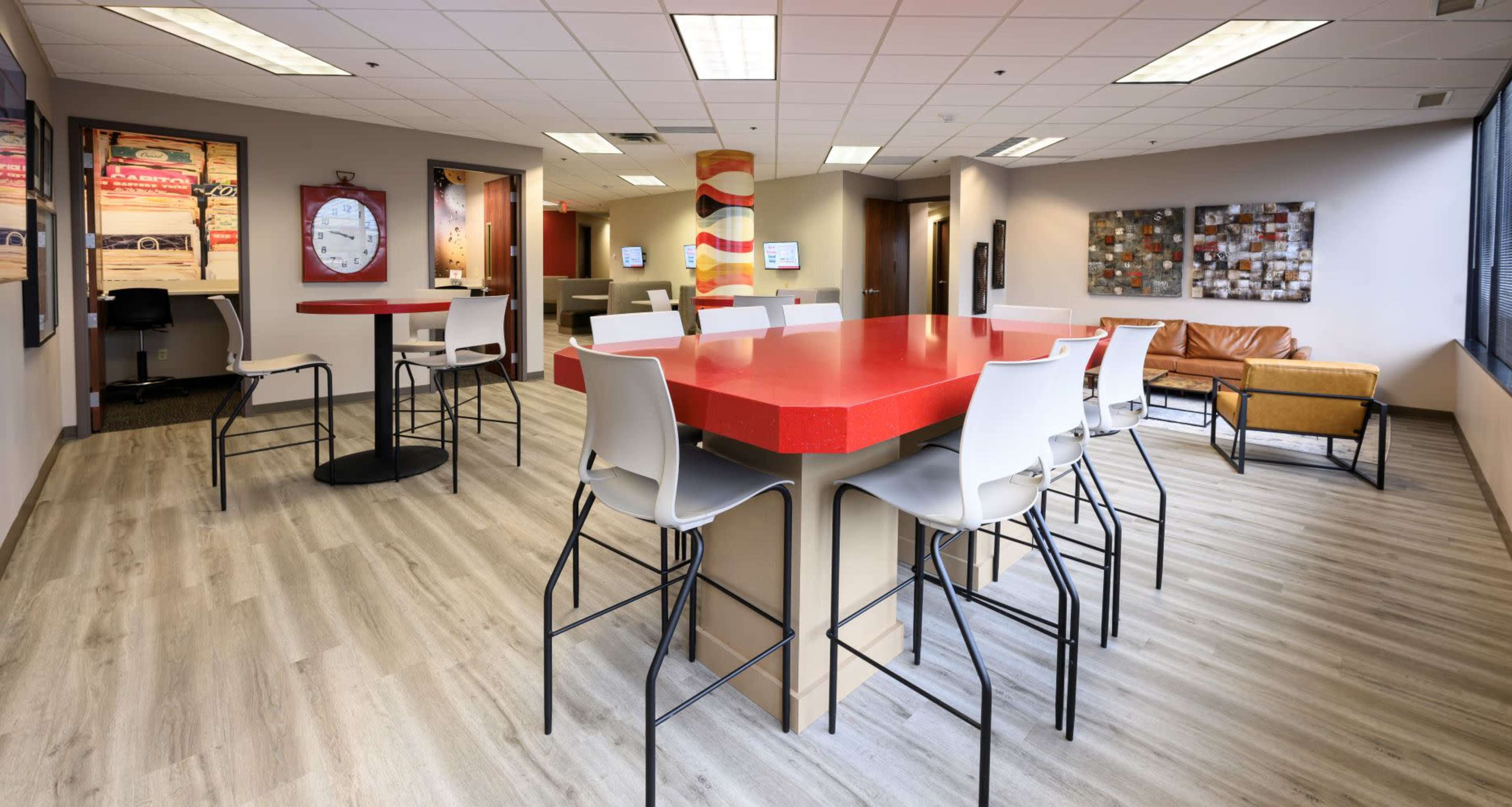 The image shows a modern office lounge featuring a red table with high stools, a leather couch, and various wall decorations.