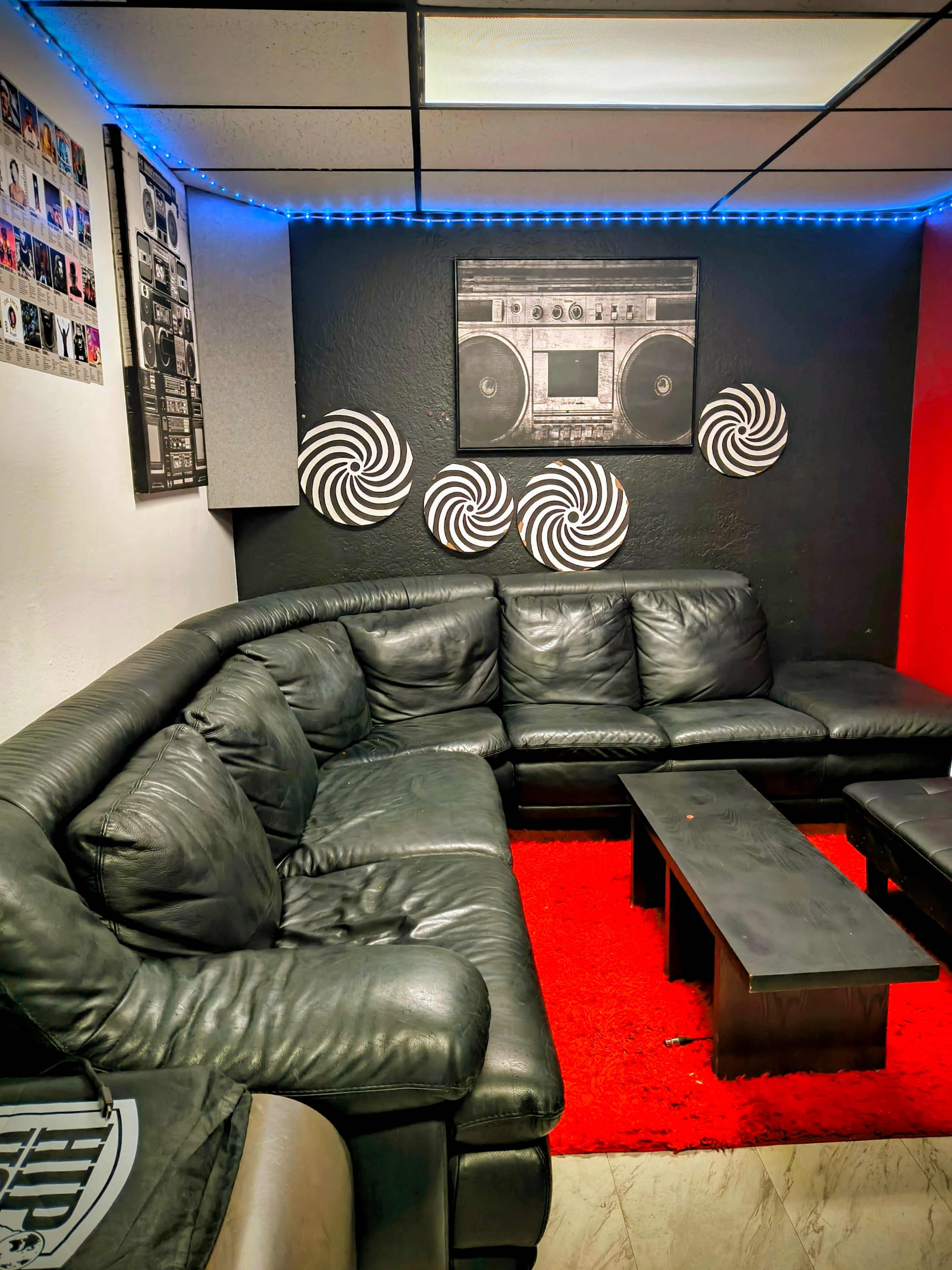 The image shows a modern lounge area with a large black sectional sofa, a low coffee table, and walls adorned with decorative elements, including a boombox art piece and spiral patterns.