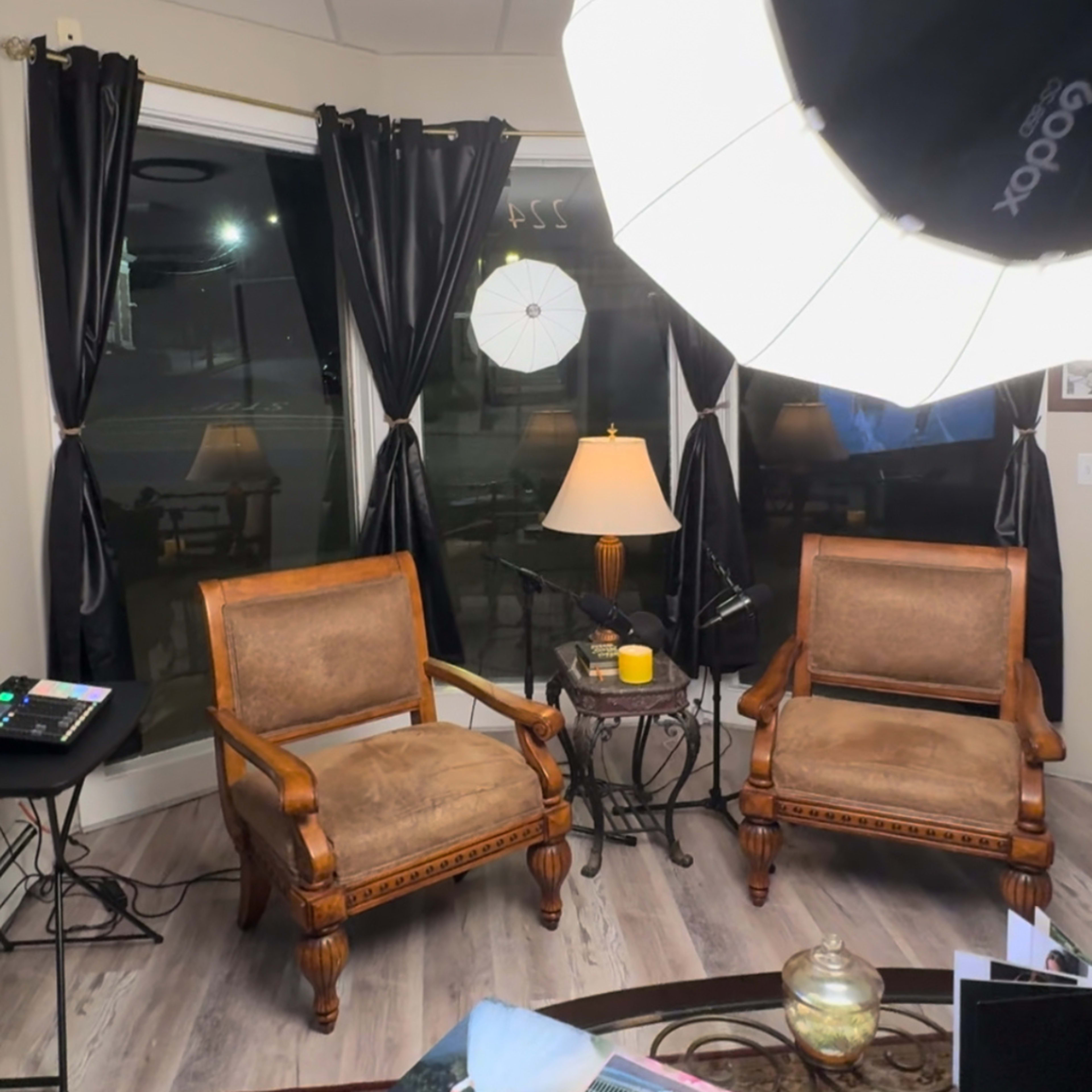 The image shows a cozy room featuring two wooden armchairs, a small round table with a lamp, and large windows covered with black curtains.