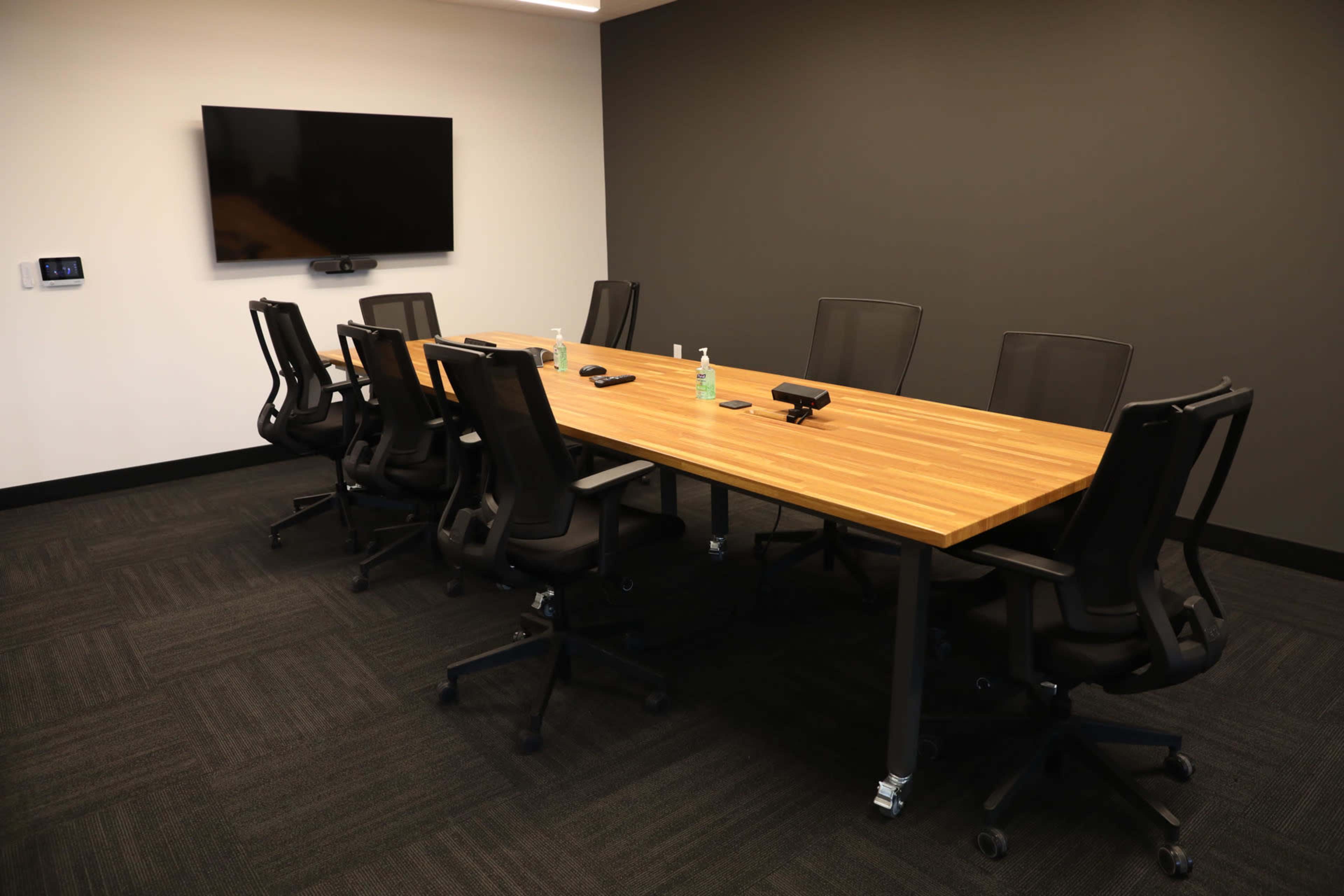 A conference room features a large wooden table surrounded by eight black office chairs, with a television mounted on the wall.