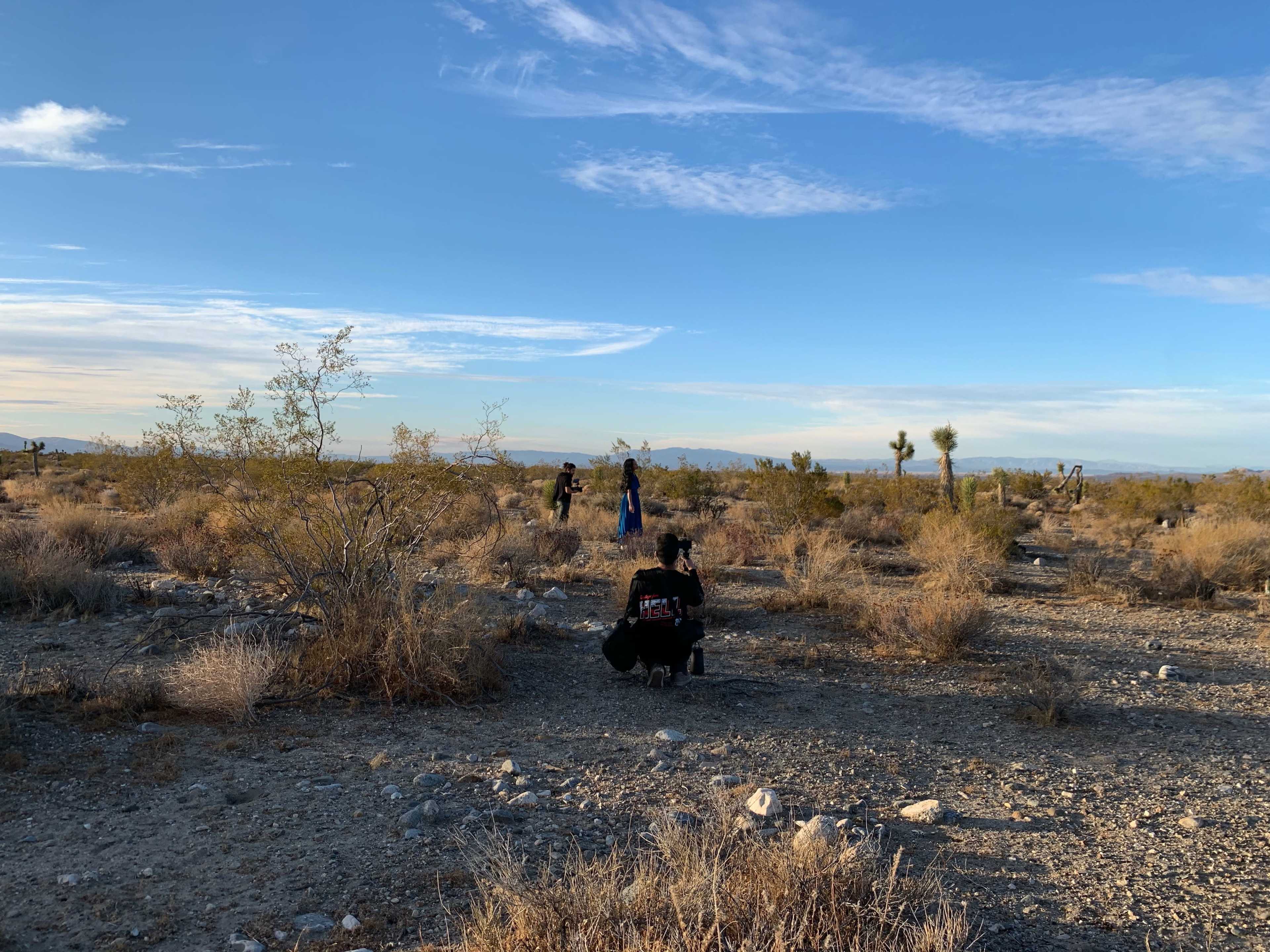 A group of people is positioned in a desert landscape, with sparse vegetation and distant mountains under a clear blue sky.