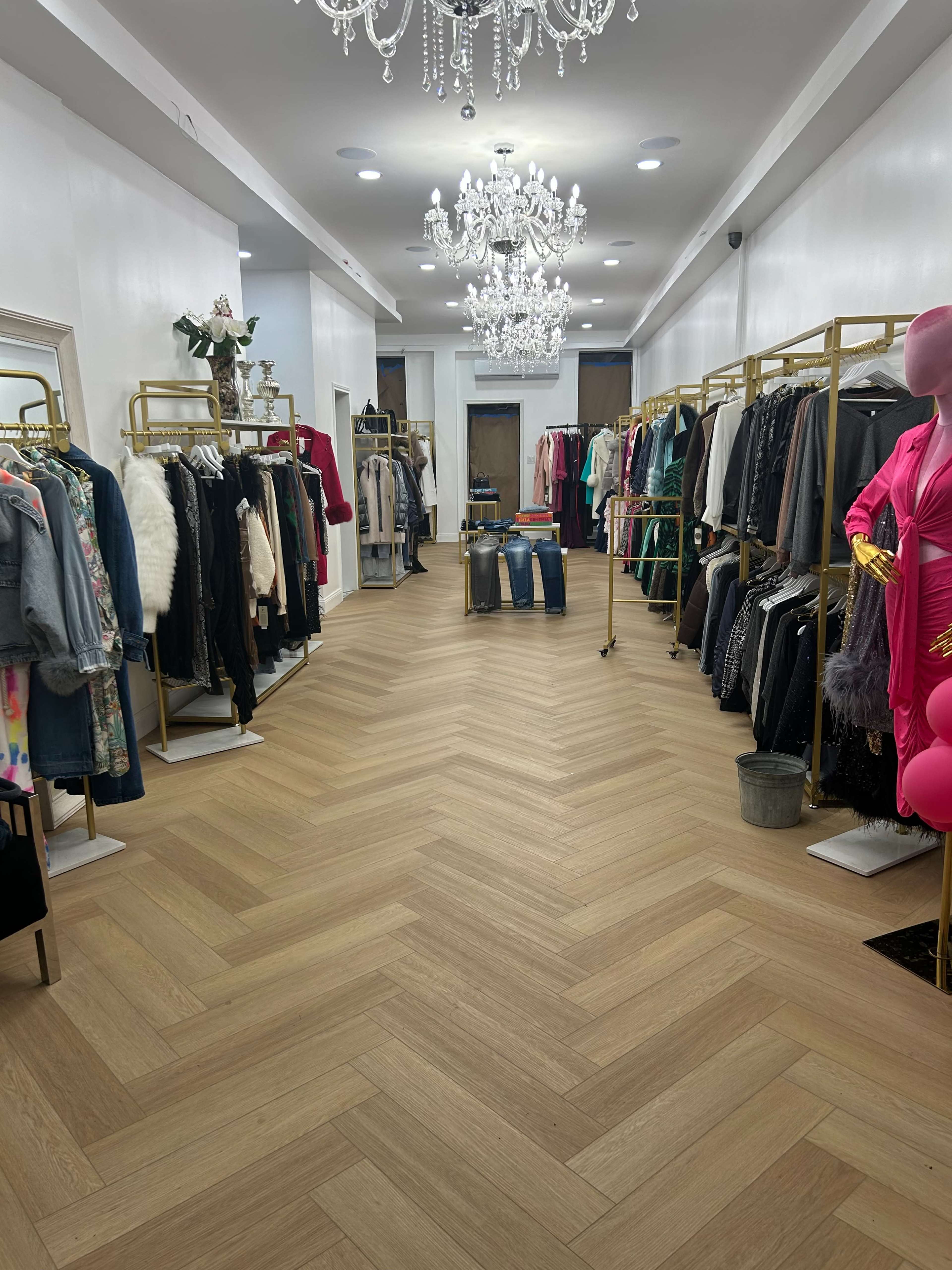 The image shows an interior of a clothing boutique featuring racks of apparel on both sides and chandeliers overhead.