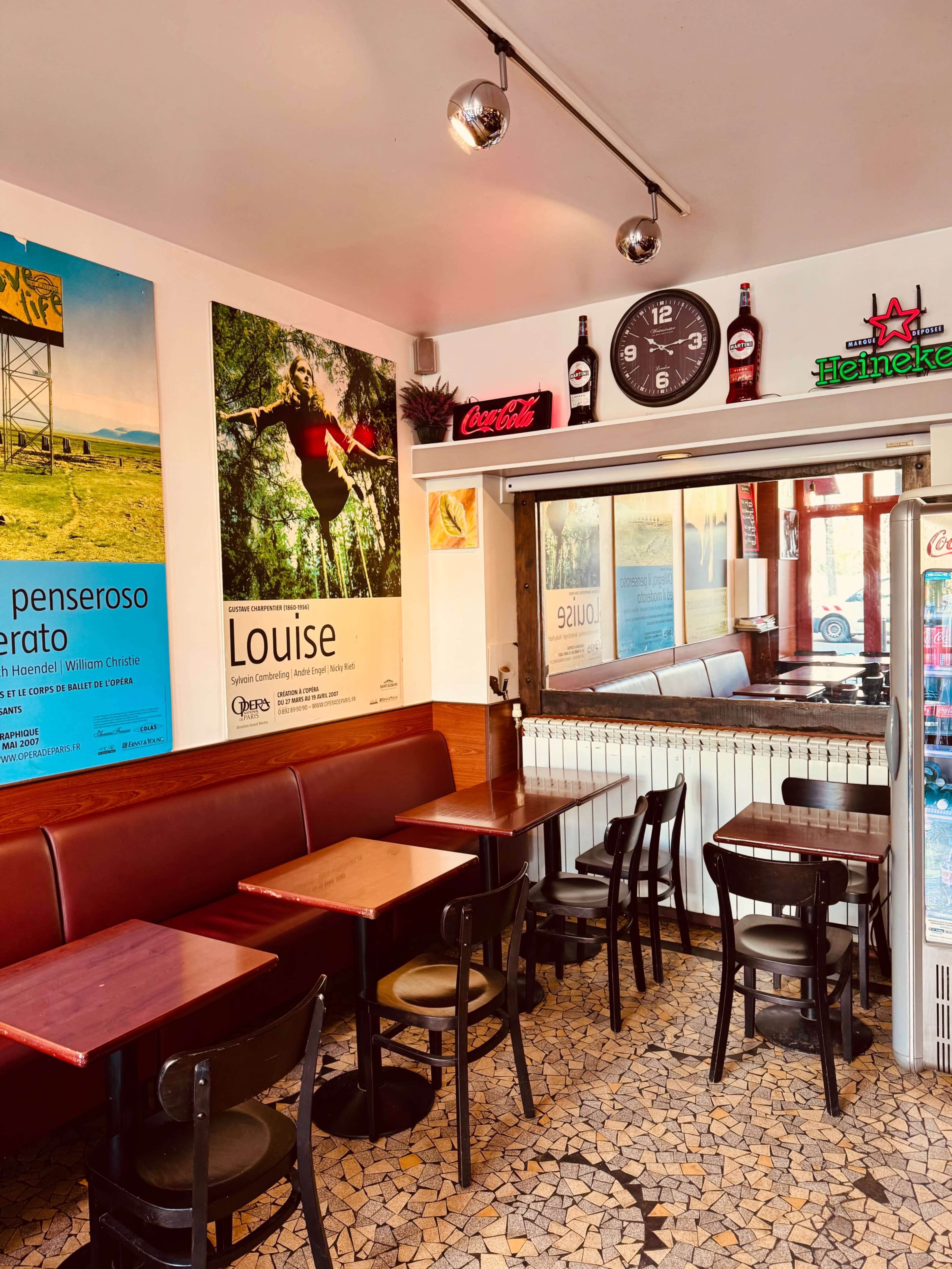 The image shows a cozy café with a tiled floor, wooden tables, and chairs, featuring colorful posters on the walls and a large clock above a mirrored bar area.