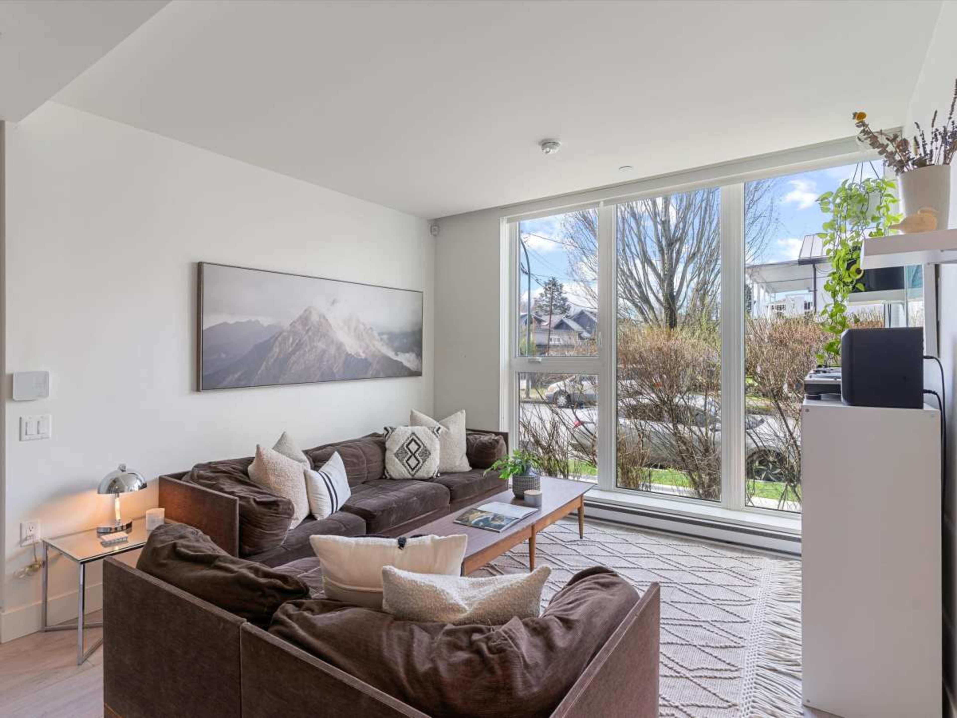 The image shows a modern living room featuring a large window, a sectional sofa with decorative pillows, and a mountain landscape photograph on the wall.