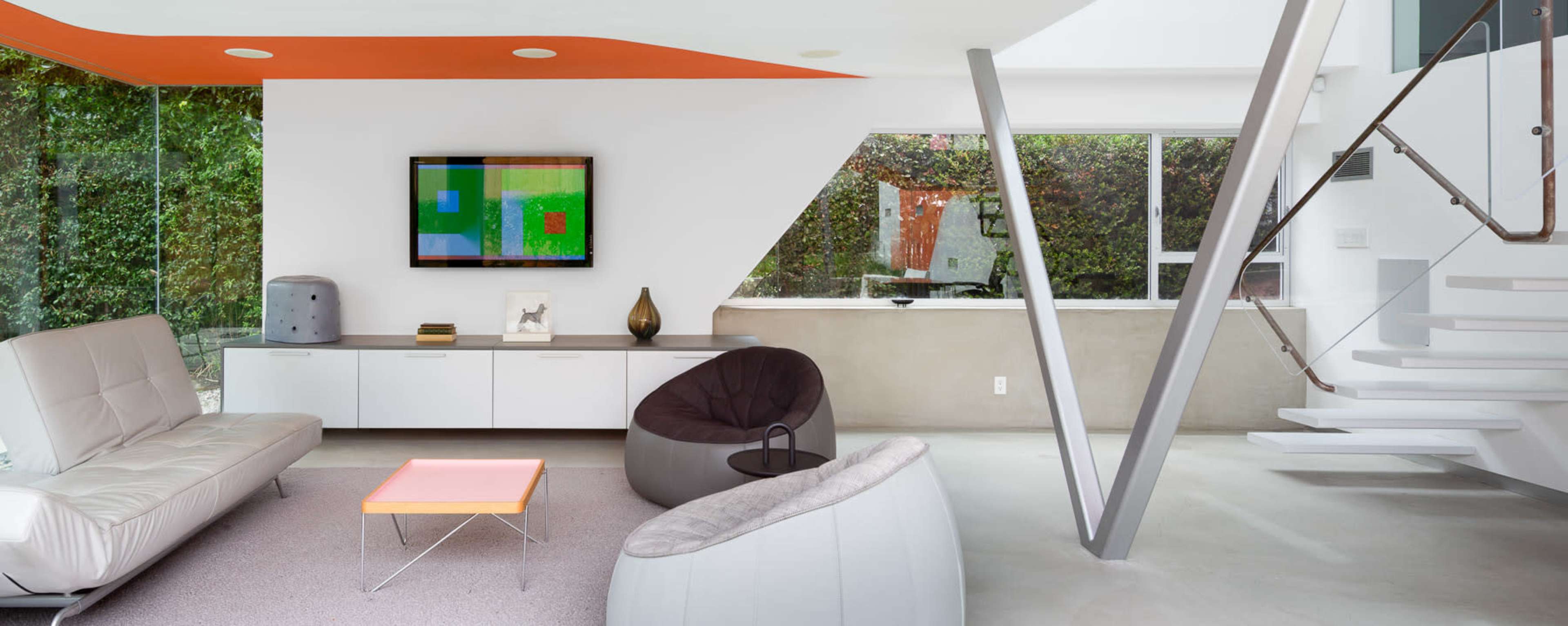 The room features modern furniture, a large window with greenery outside, and a staircase with a support beam.