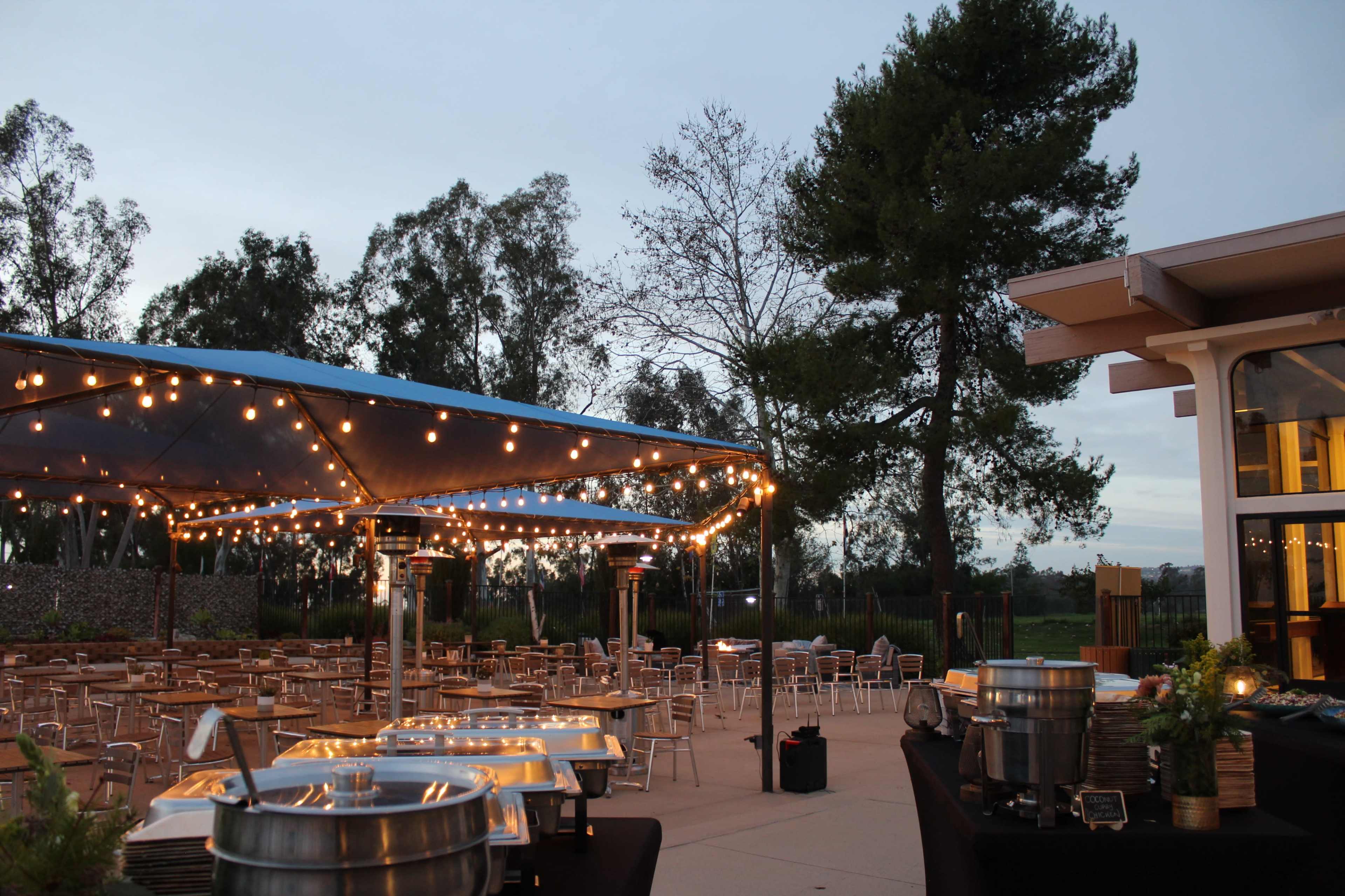 The scene features an outdoor catering setup with a large tent adorned with string lights and tables arranged under a blue sky at dusk.