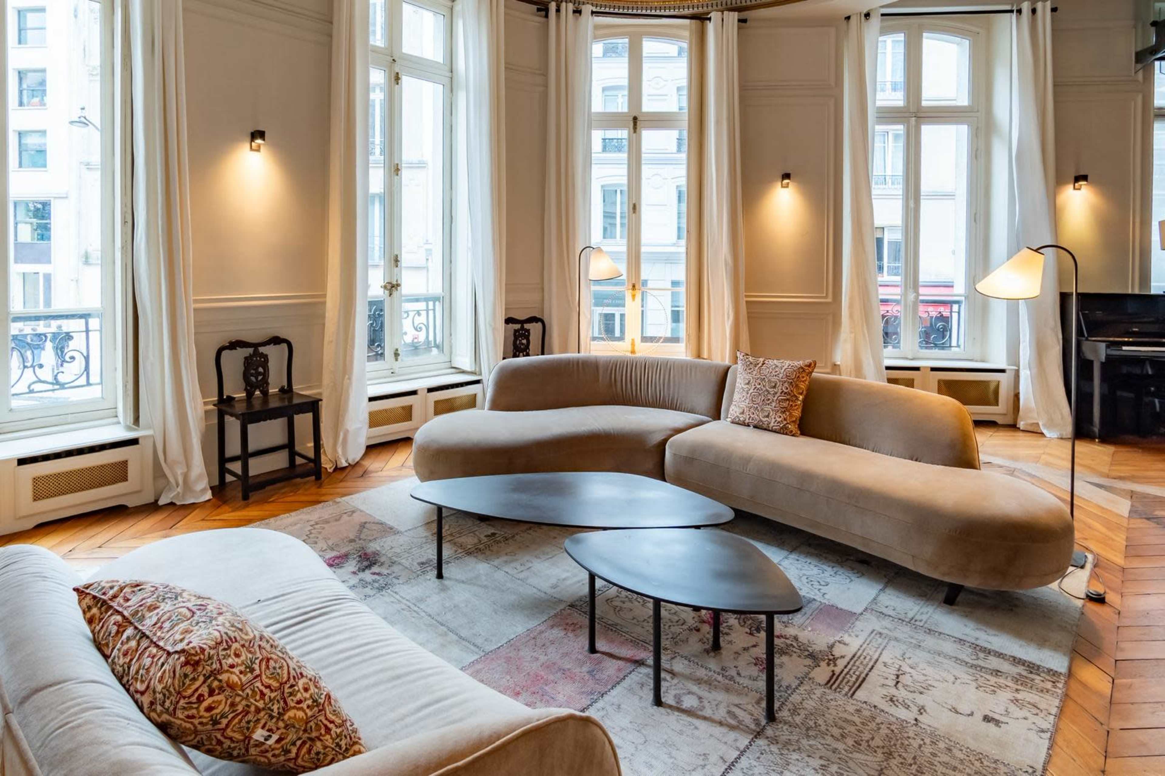 The image shows a modern, elegantly designed living room with curved couches, a patterned area rug, and large windows that let in natural light.
