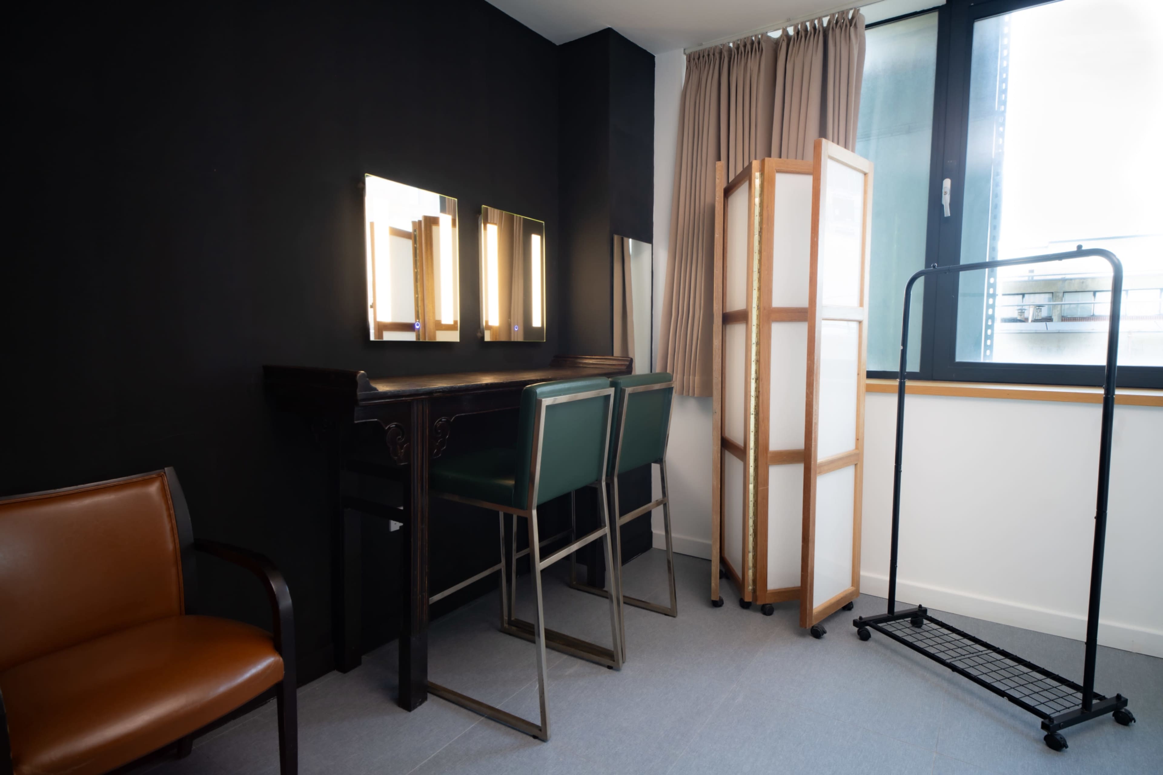 The image shows a small room featuring two green chairs at a vanity table with three vertical lights, a folding screen, and a clothing rack.