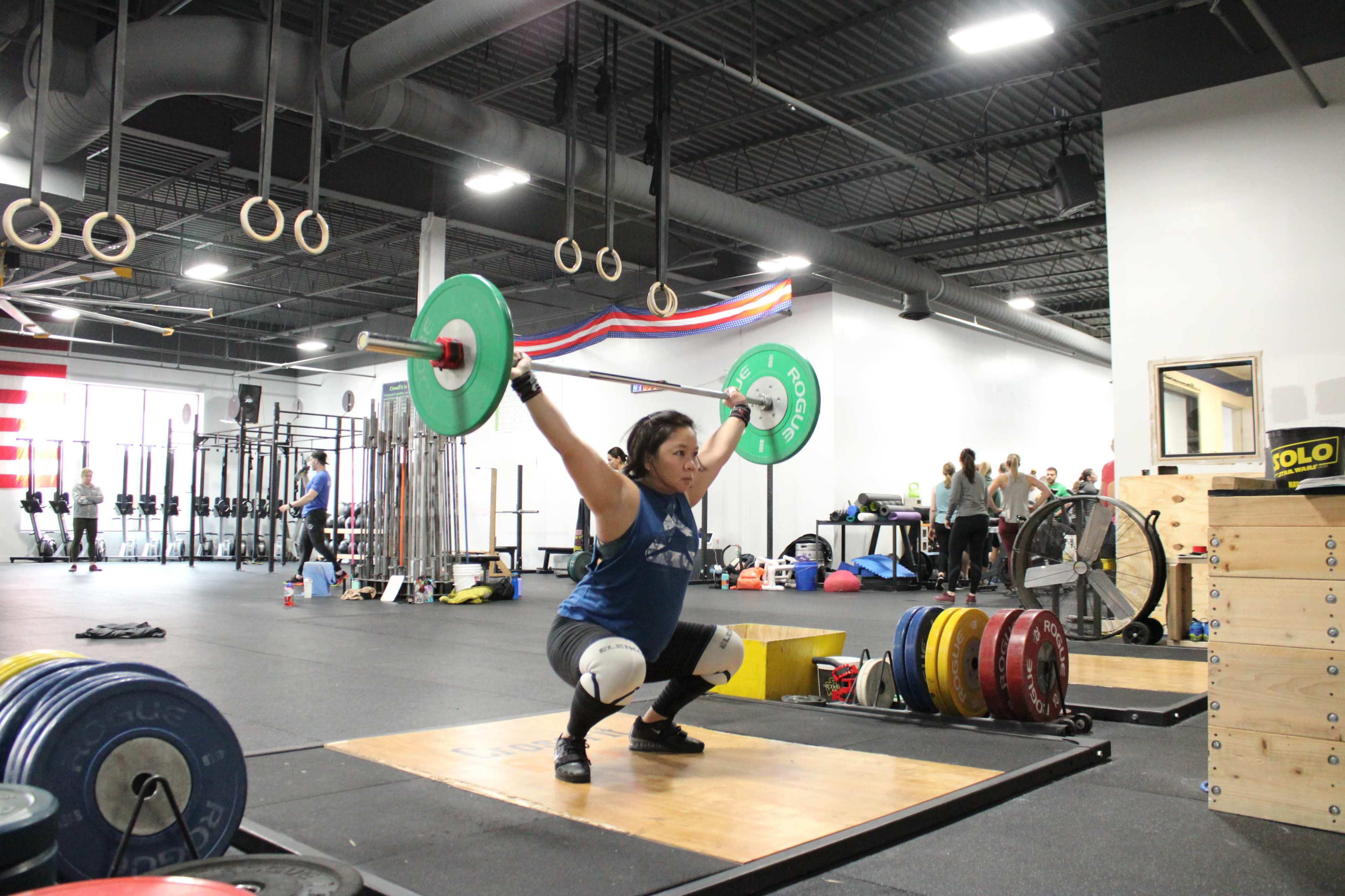 A person performs an overhead lift with a barbell in a gym filled with weightlifting equipment and mats.