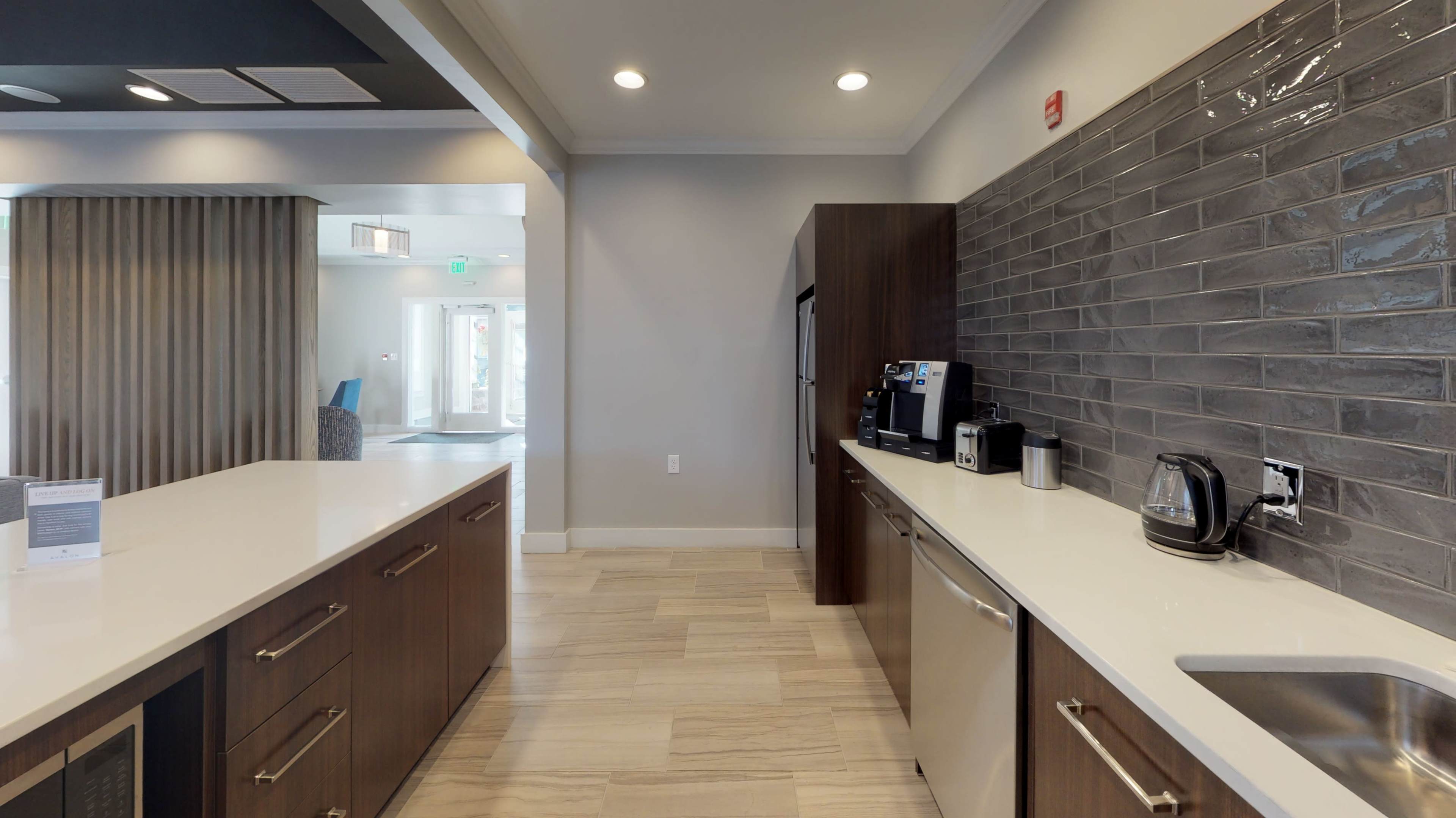 A modern kitchen area features wooden cabinetry, a stainless steel refrigerator, and a countertop with a coffee maker and kettle, set against a gray tiled backsplash.