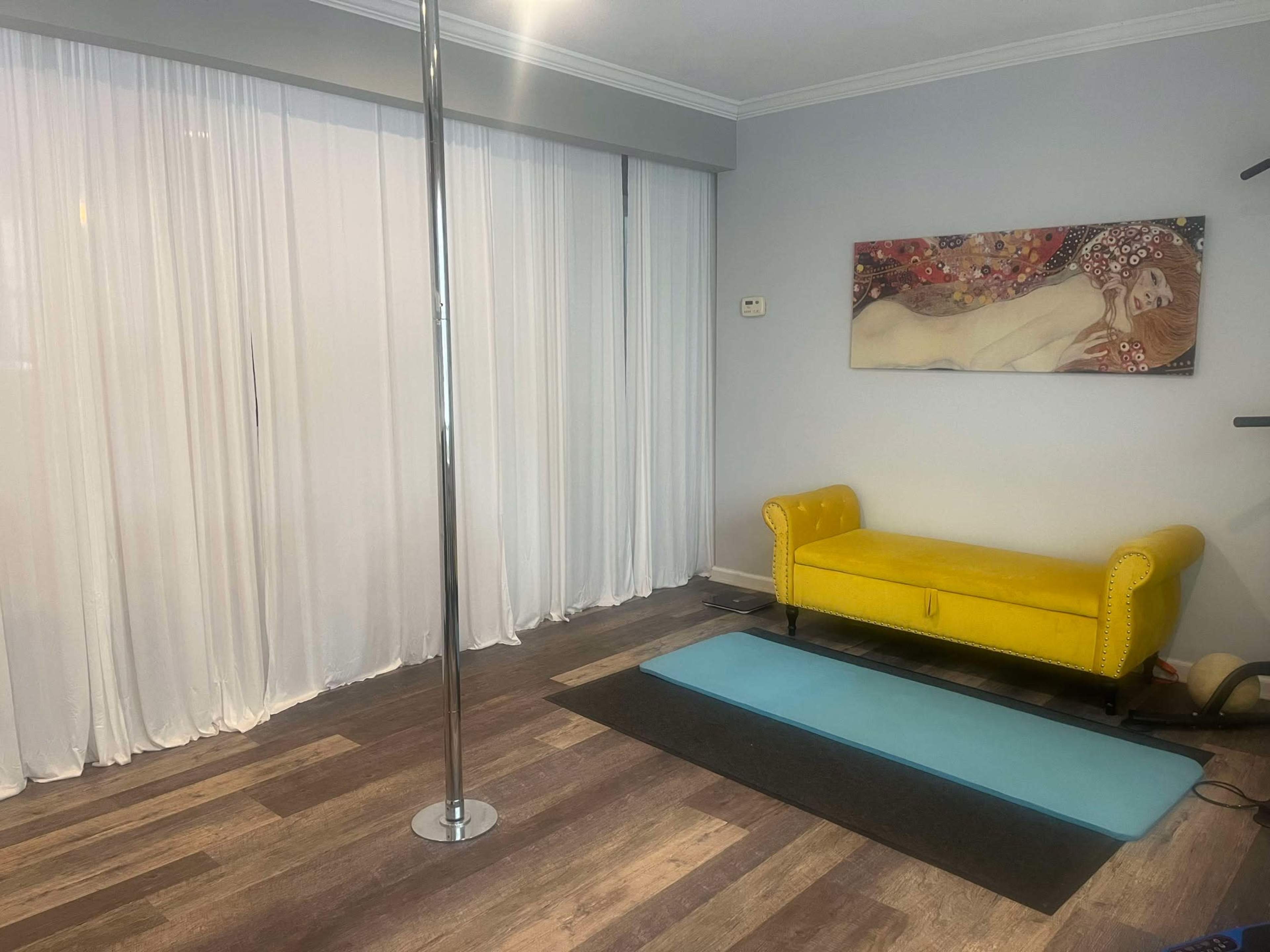 The image shows a room with a pole, a yellow couch, and a blue exercise mat on a wooden floor, with sheer white curtains covering a window.