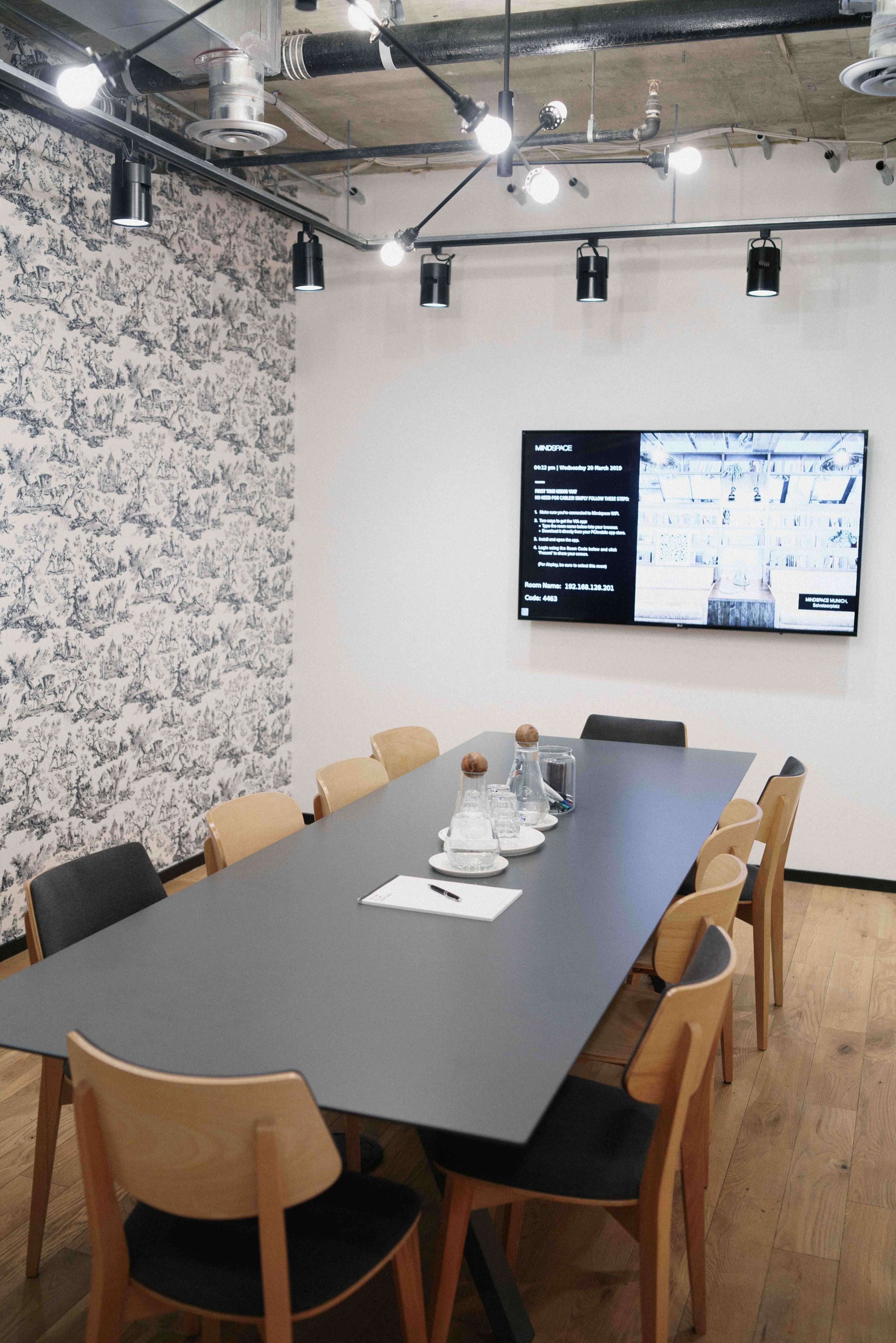 A modern conference room features a long dark table surrounded by wooden chairs, with a large screen displaying information on one wall.