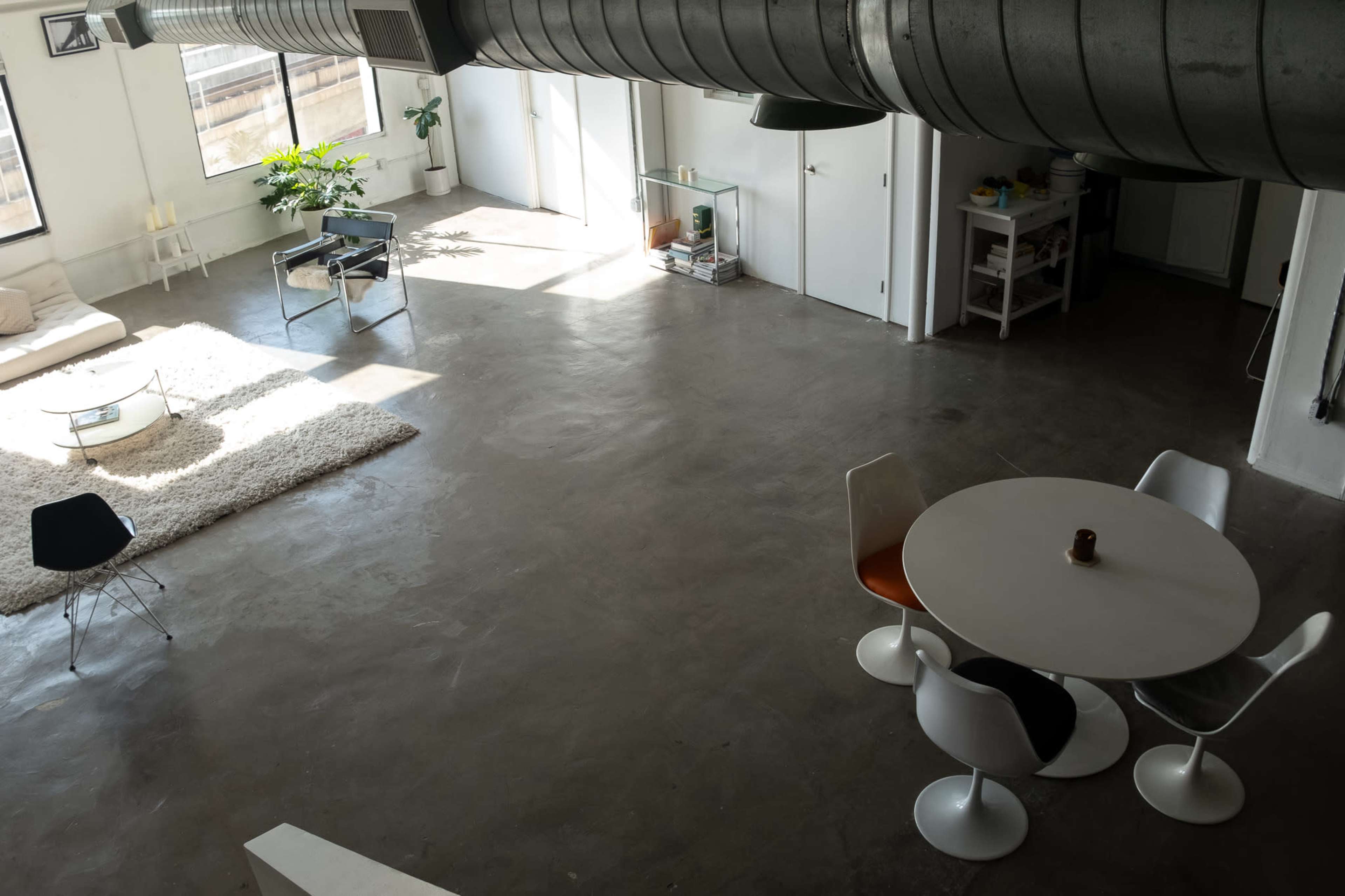 The image shows a spacious, minimalist interior with a living area featuring a sofa and a rug, and a dining area with a white circular table and chairs.