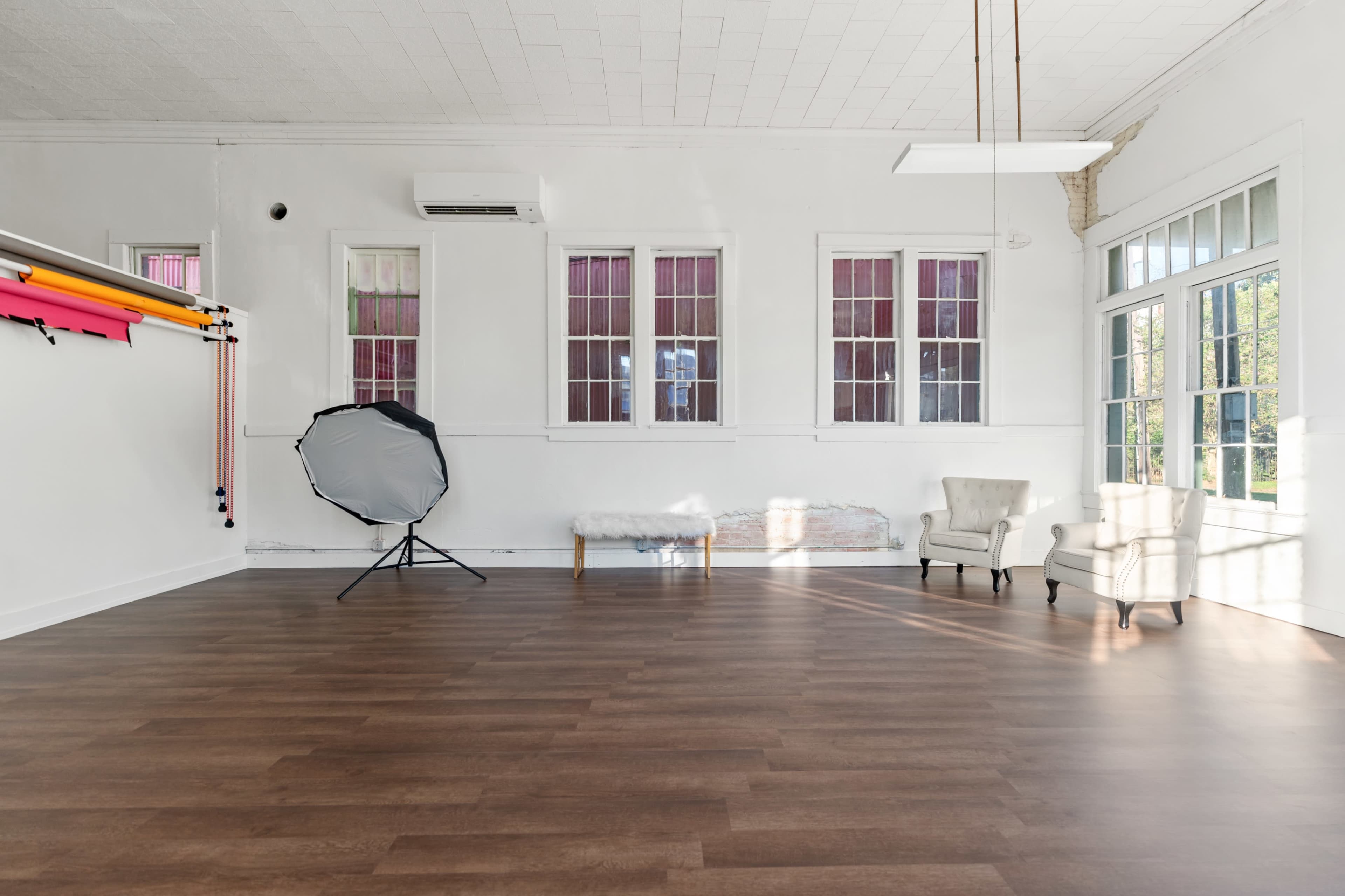The image shows a spacious, well-lit room with wooden flooring, featuring a photography backdrop setup on the left and a chair beside a window on the right.