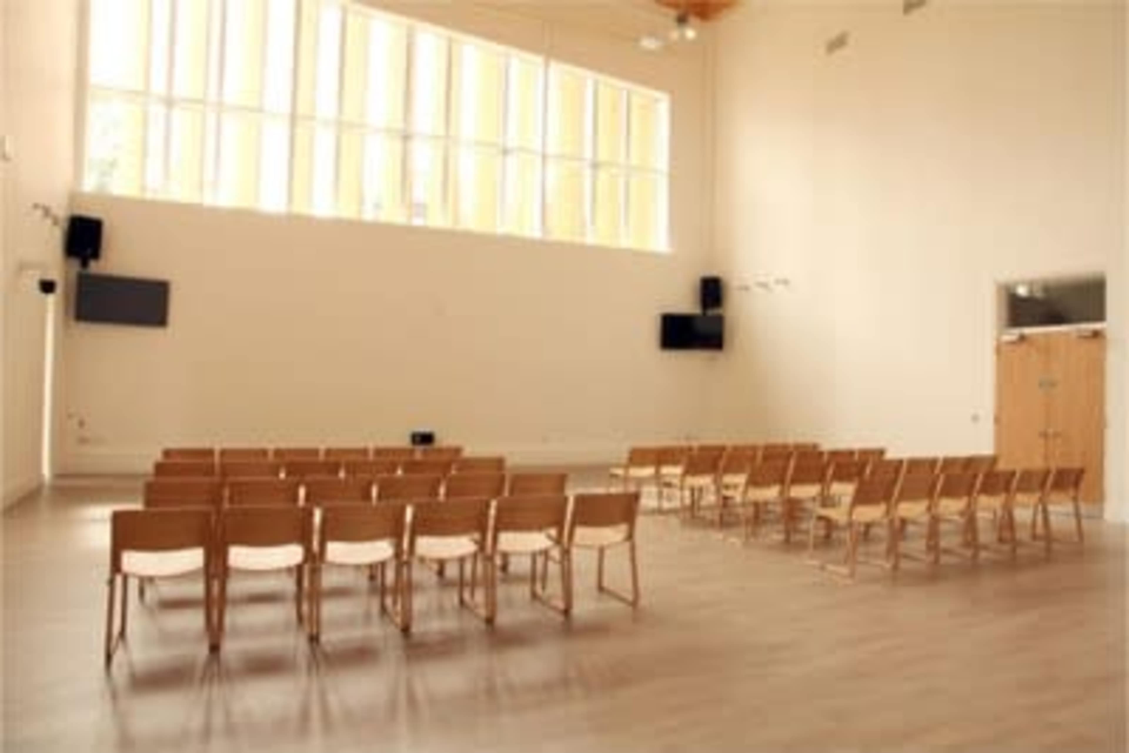 A spacious room with wooden chairs arranged in rows, featuring large windows and audio-visual equipment on the walls.