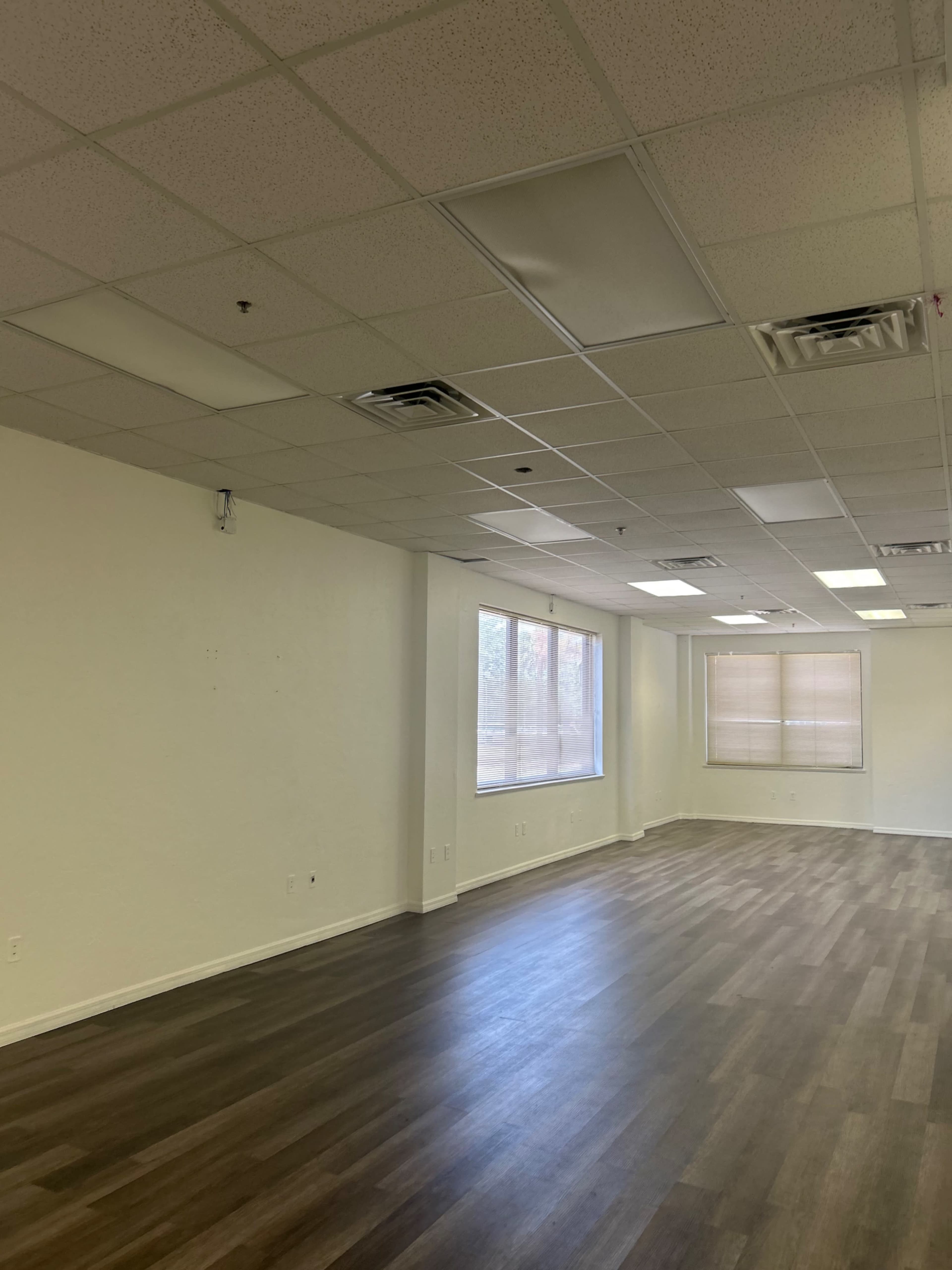 The image shows an empty room with light-colored walls, large windows, and a wooden floor.