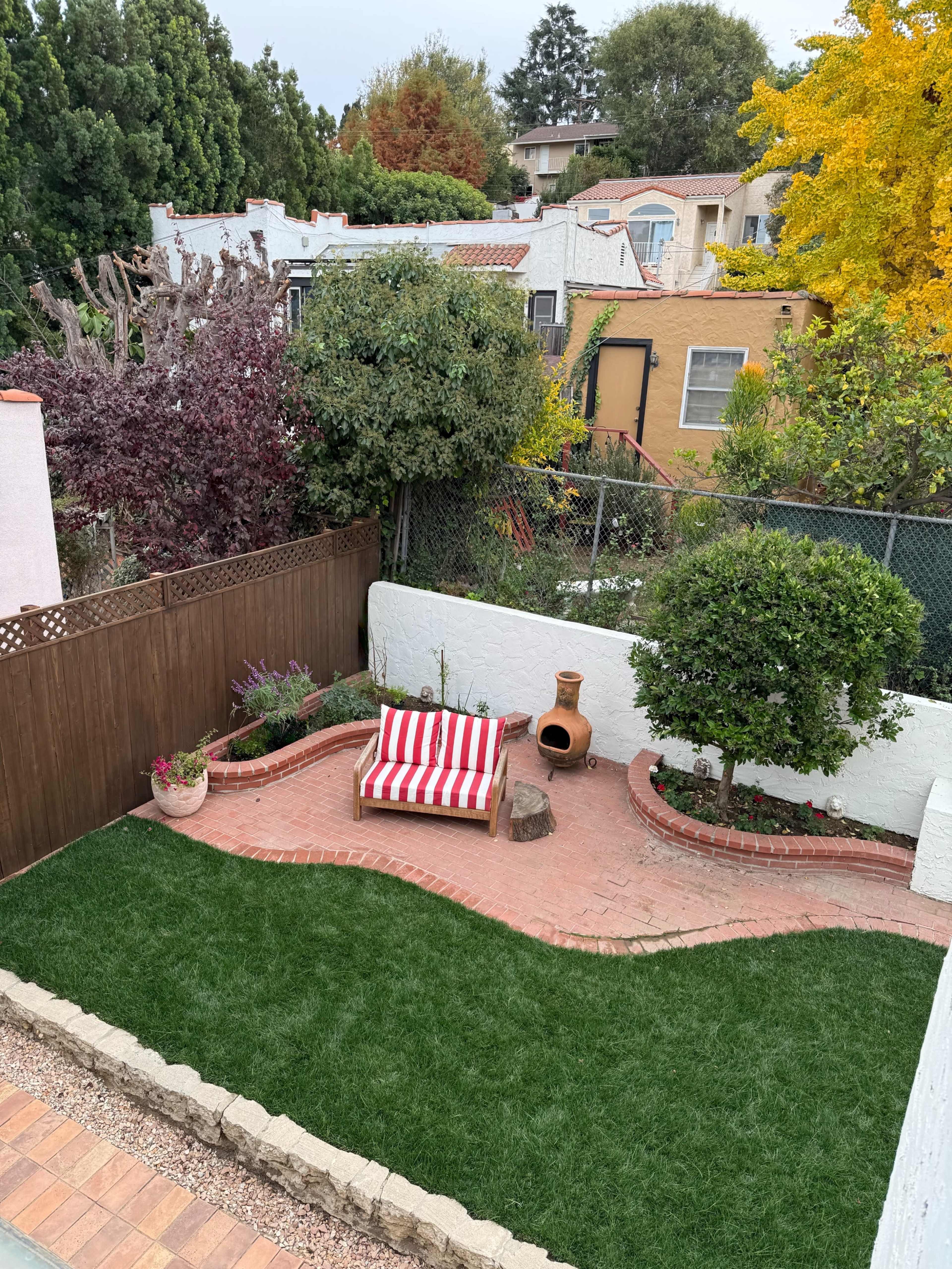 The image shows a small outdoor garden area with a brick patio, a striped lounge chair, a decorative stone, and greenery surrounded by a wooden fence.