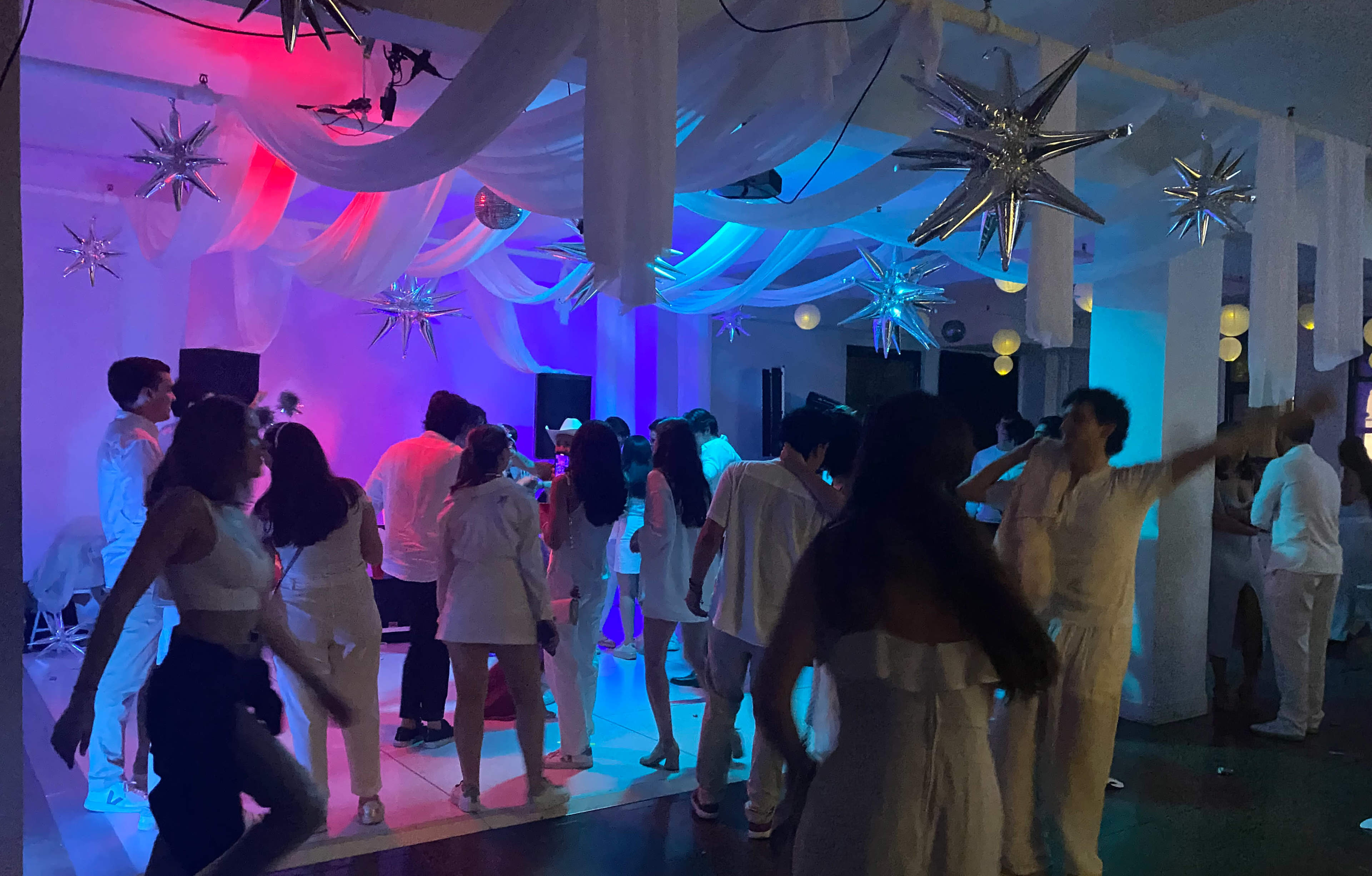 A group of people dressed in white are dancing at an event illuminated by colorful lights with decorative stars hanging from the ceiling.