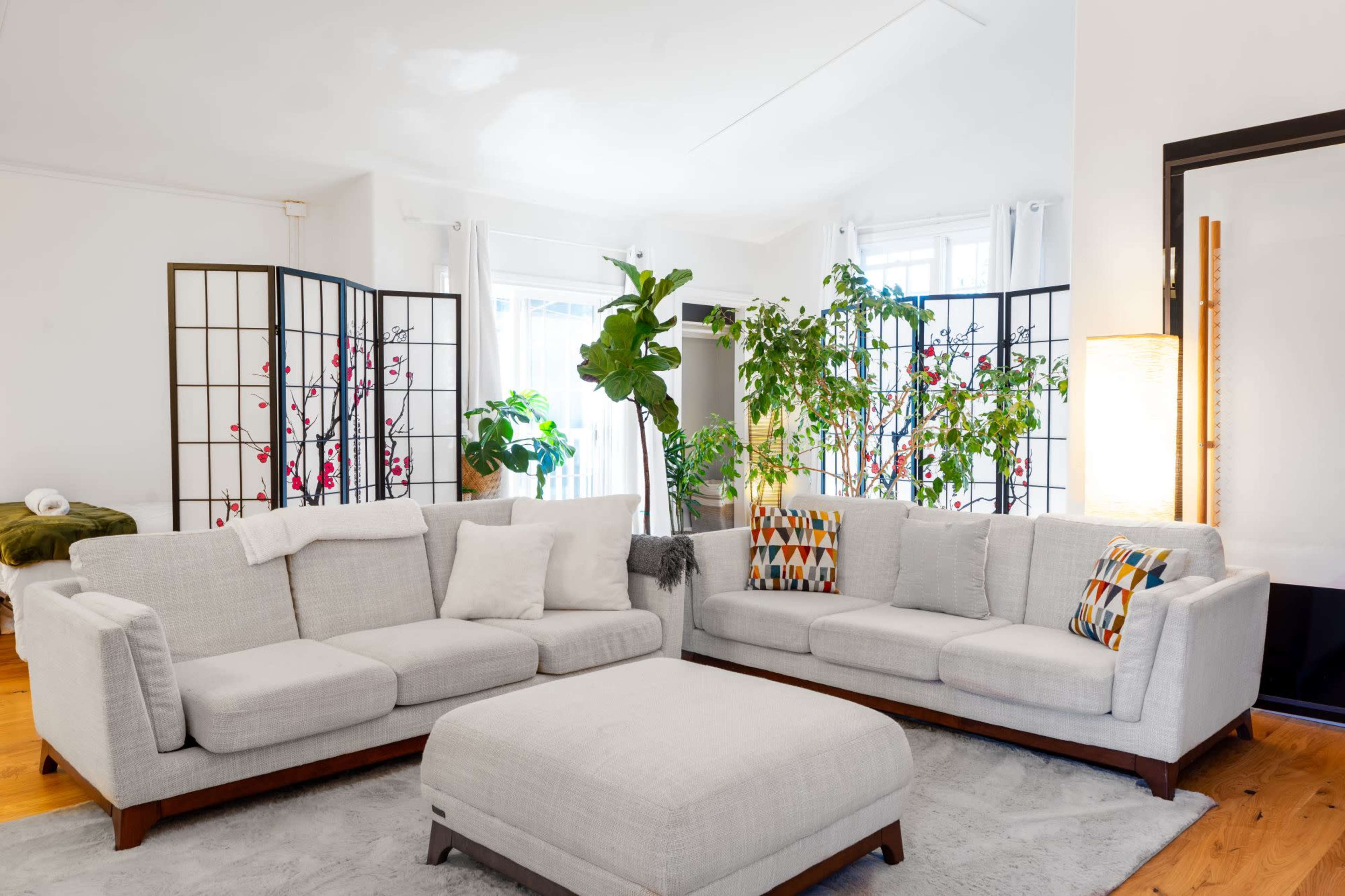 A light-colored living room features a spacious sofa, an ottoman, and decorative plants arranged behind a folding screen.