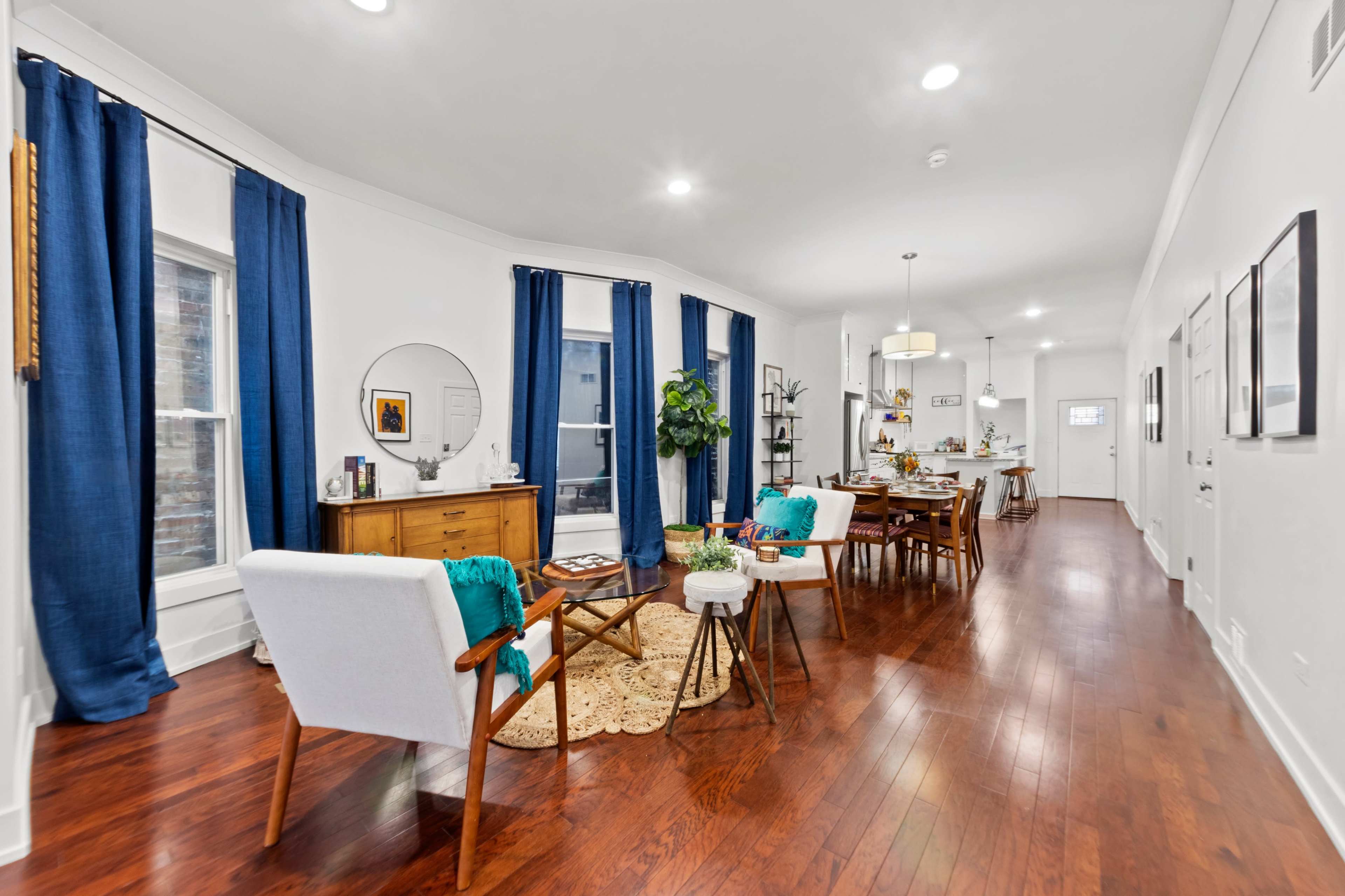 A modern living space features hardwood floors, white walls, and a combination of a dining area and lounge with blue curtains, wooden furniture, and decorative plants.