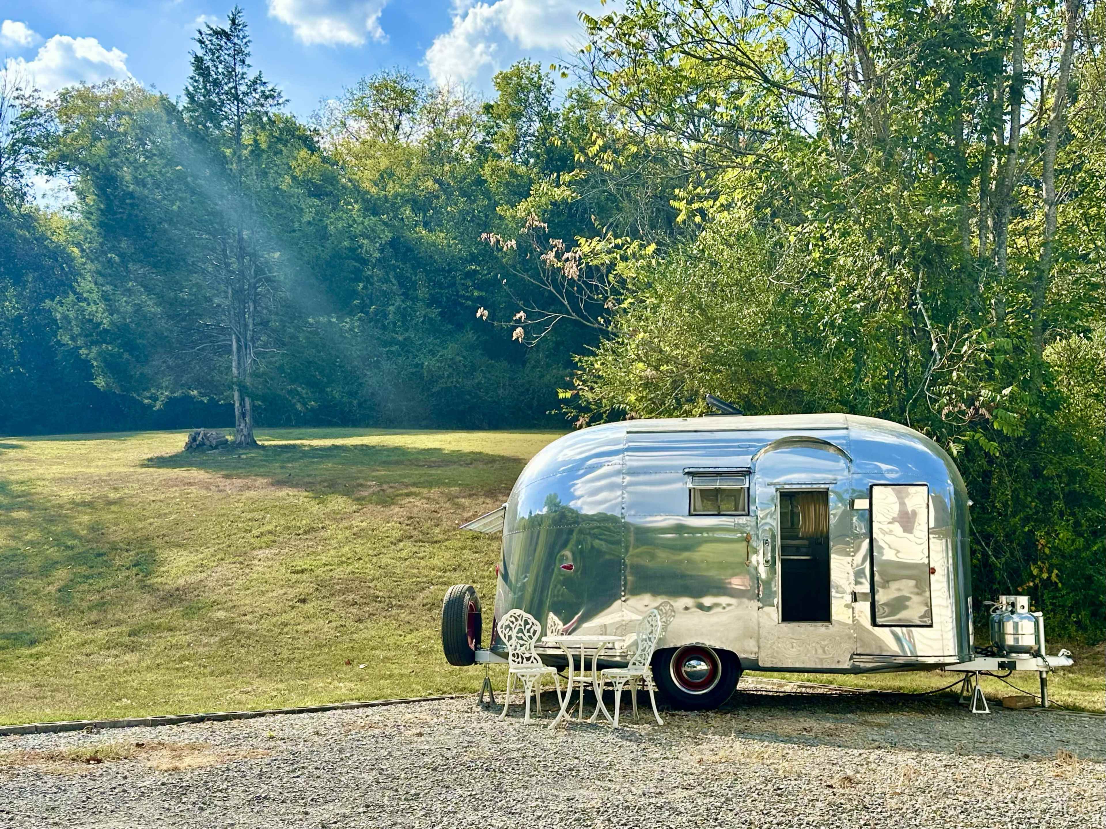 A shiny, silver Airstream trailer is parked on a gravel area, accompanied by a small white table and chairs, with a grassy field and trees in the background.