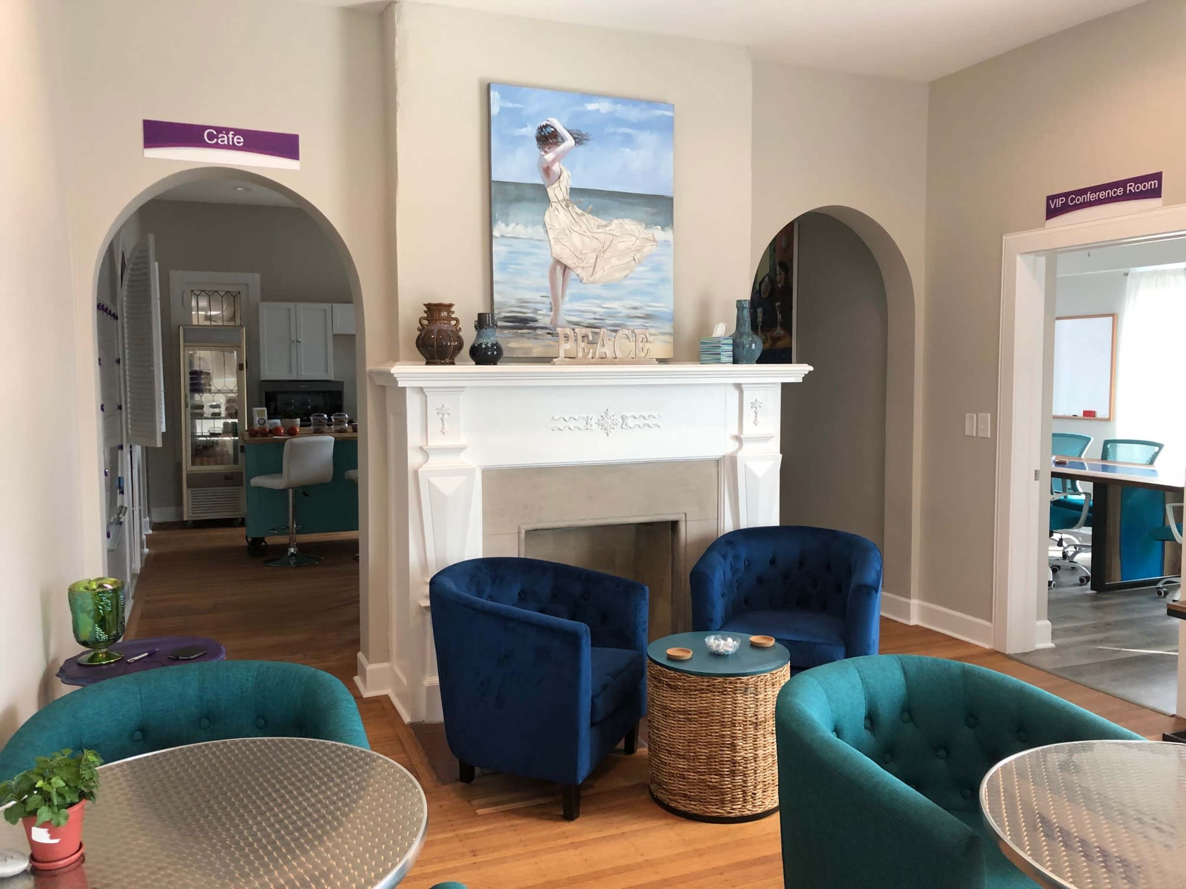 A lounge area featuring two blue upholstered chairs, a coffee table, and a painting of a woman by the ocean above a white fireplace.