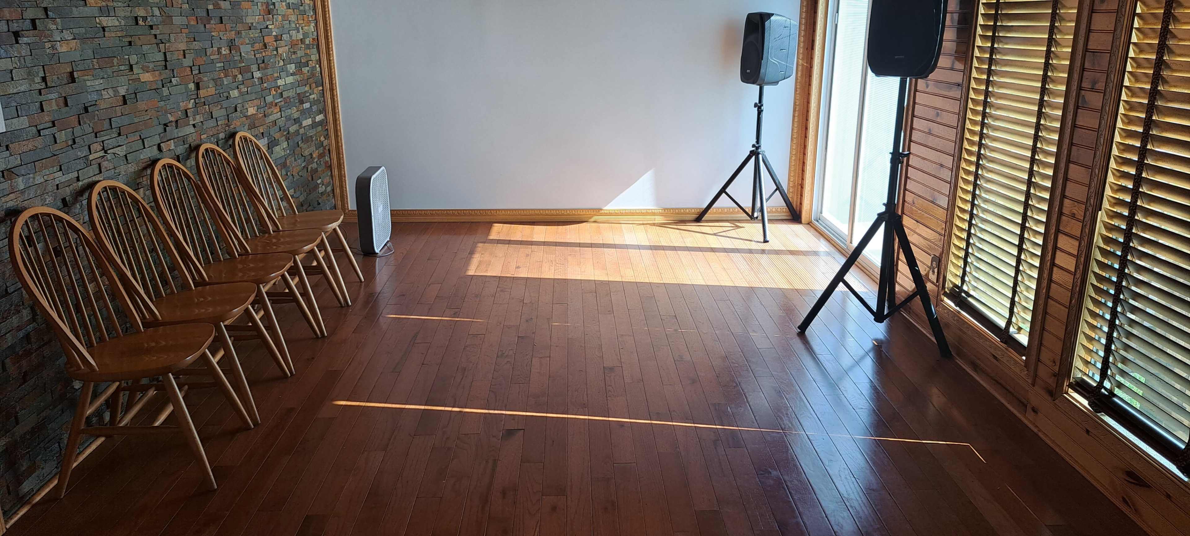 A room with wooden flooring, a stone accent wall, two speakers, a space heater, and four wooden chairs arranged in a line near a window with blinds.