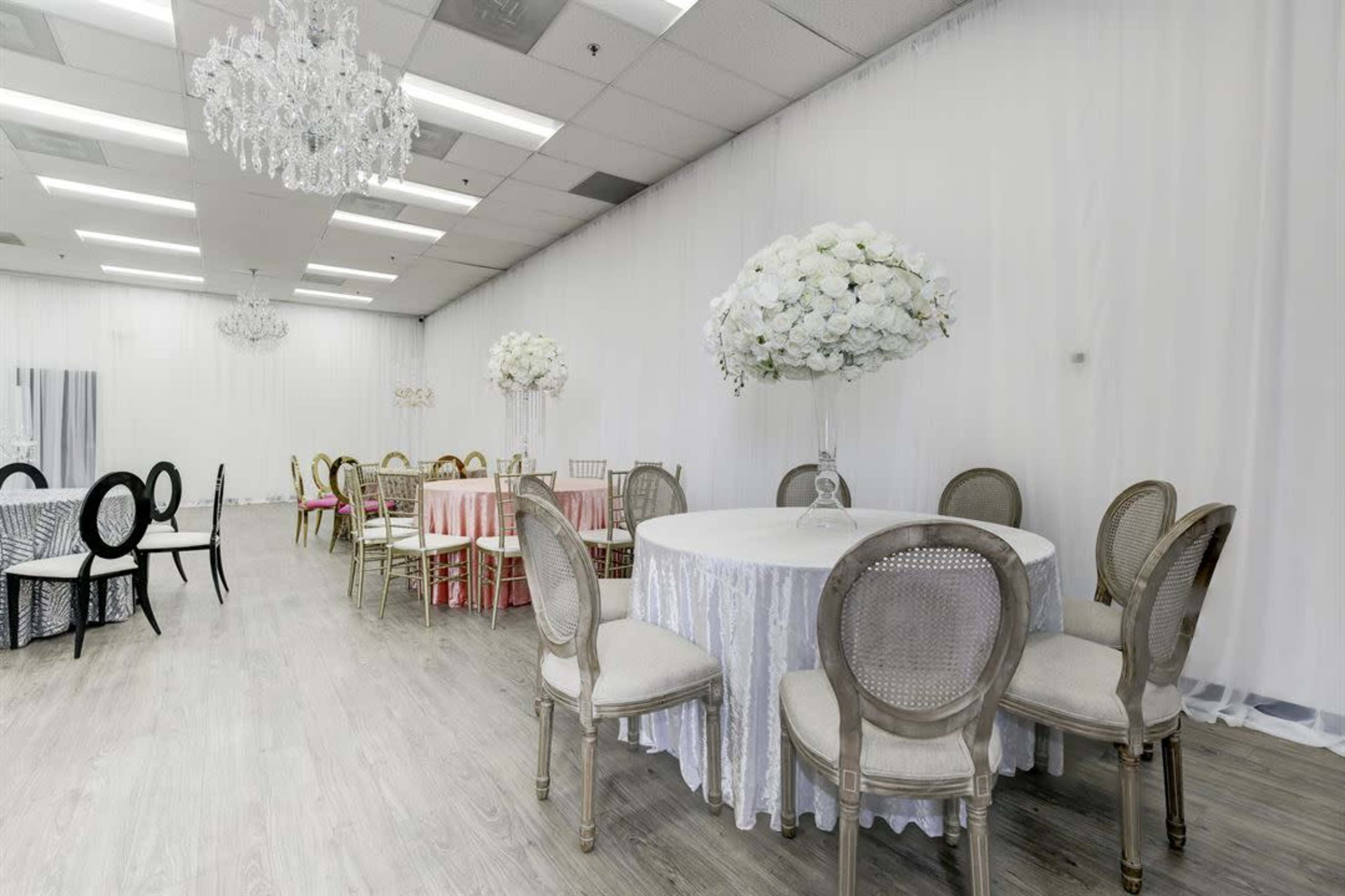 A spacious event venue features elegantly arranged round and rectangular tables adorned with floral centerpieces against a backdrop of sheer white drapes.