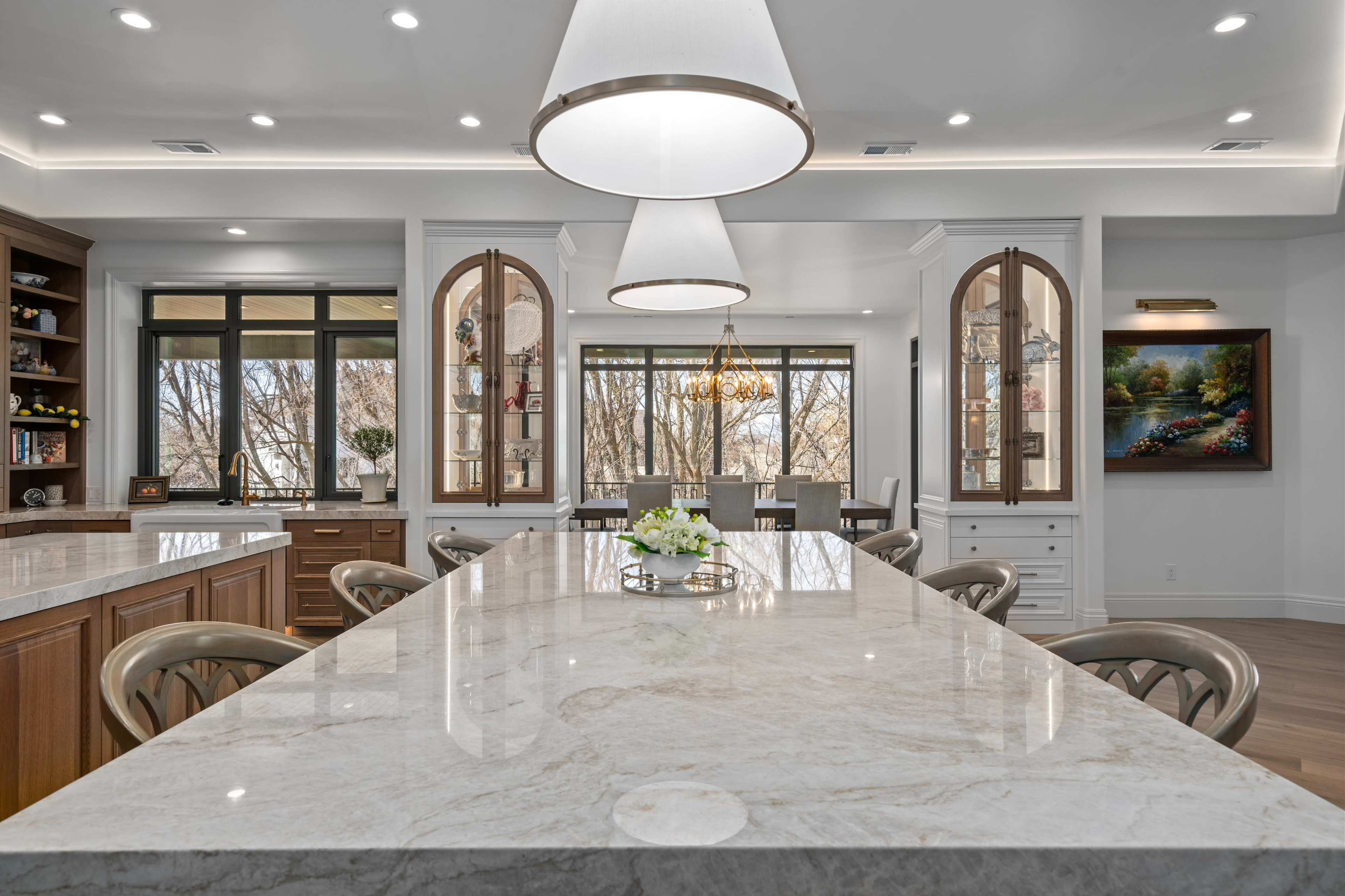 The image shows a modern kitchen with a large marble island, pendant lighting, and a dining area visible through large windows.