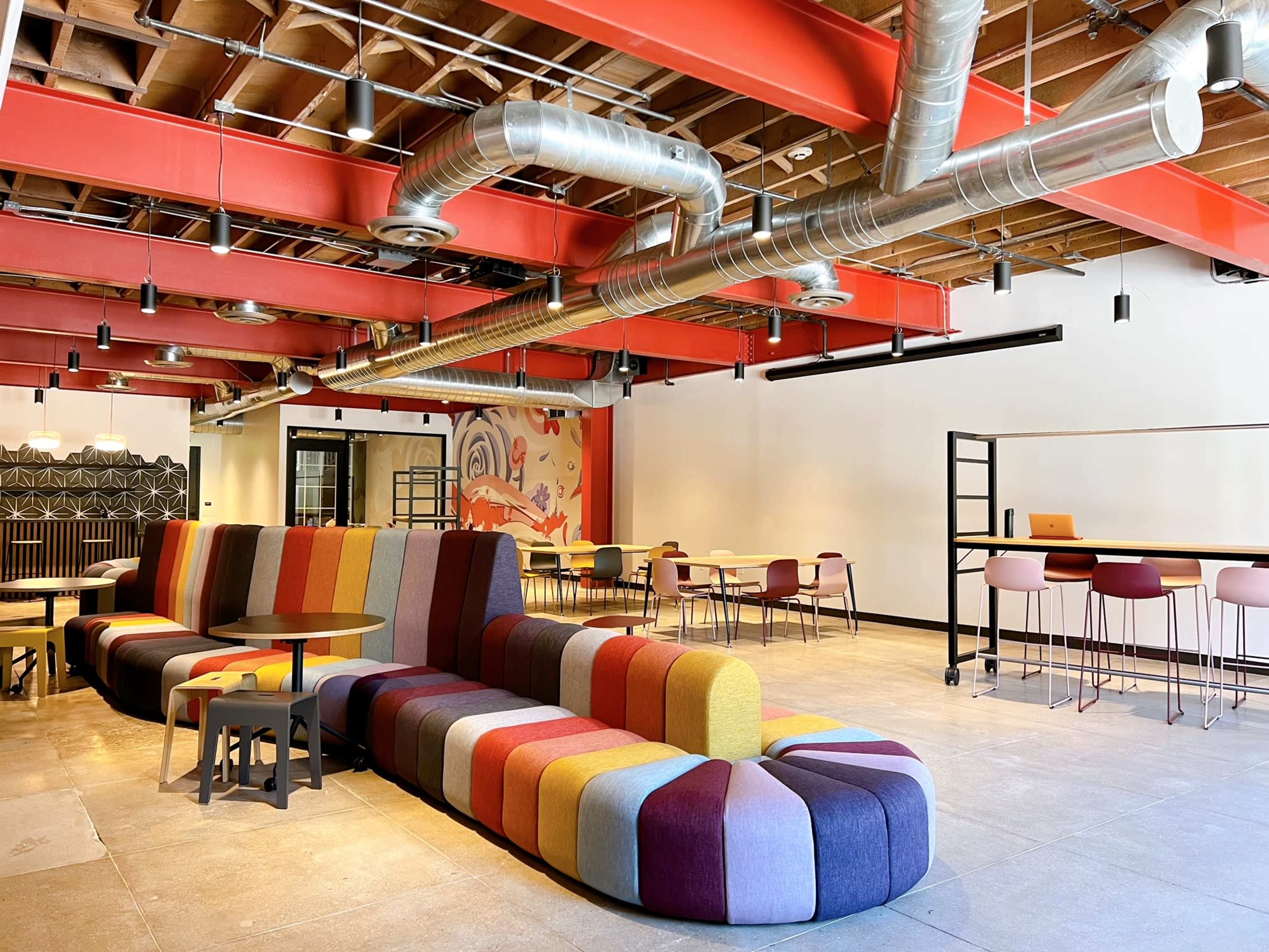 The image shows a modern lounge area featuring a colorful, curved couch, round tables, and high-top seating under exposed ductwork and beams.