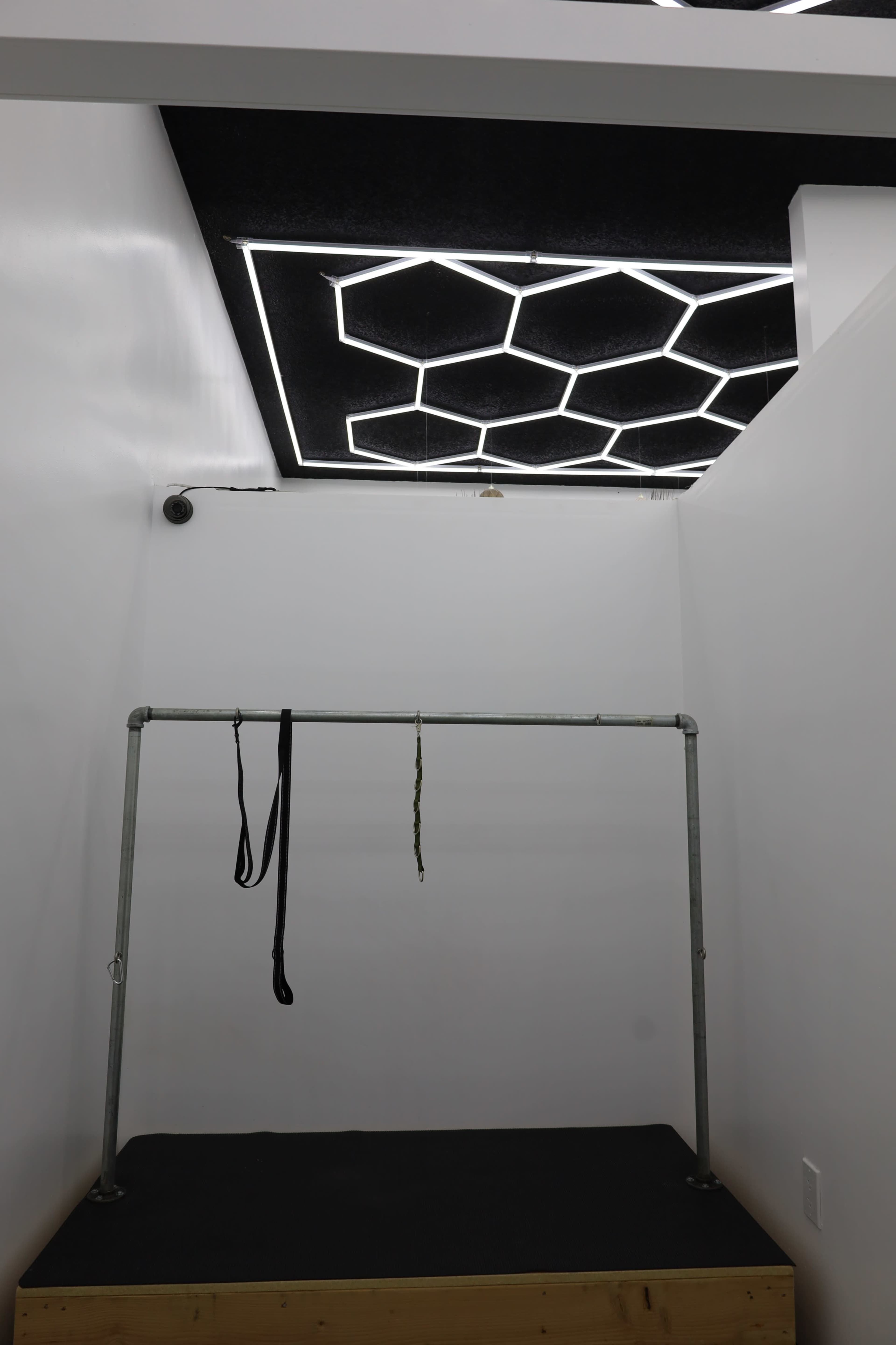 The image shows a minimalistic interior space with a framed structure hanging from the ceiling, illuminated by a geometric light fixture.