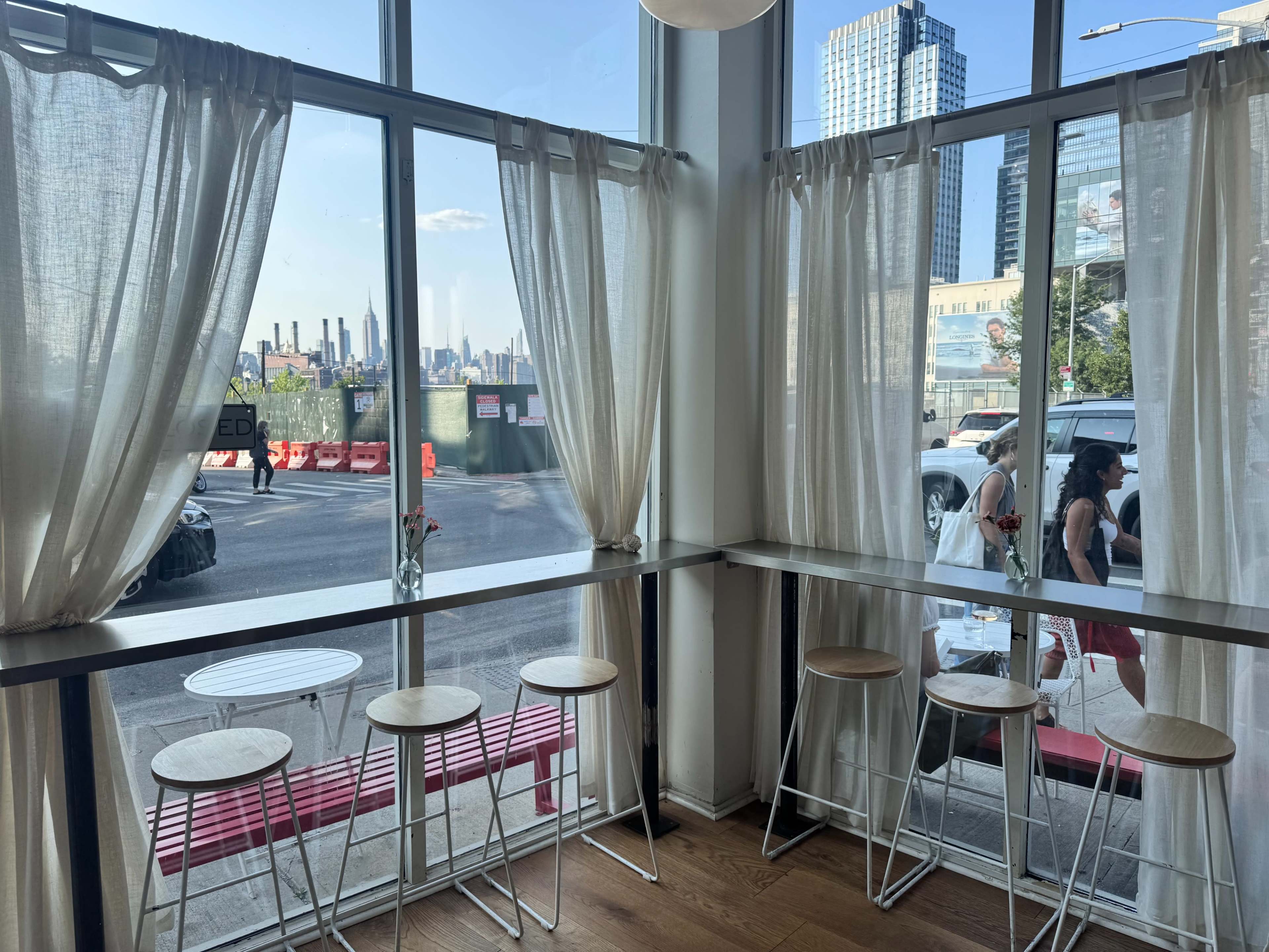 The image shows a modern café interior with a row of bar stools along a window, sheer curtains, and a view of a city street and skyline outside.