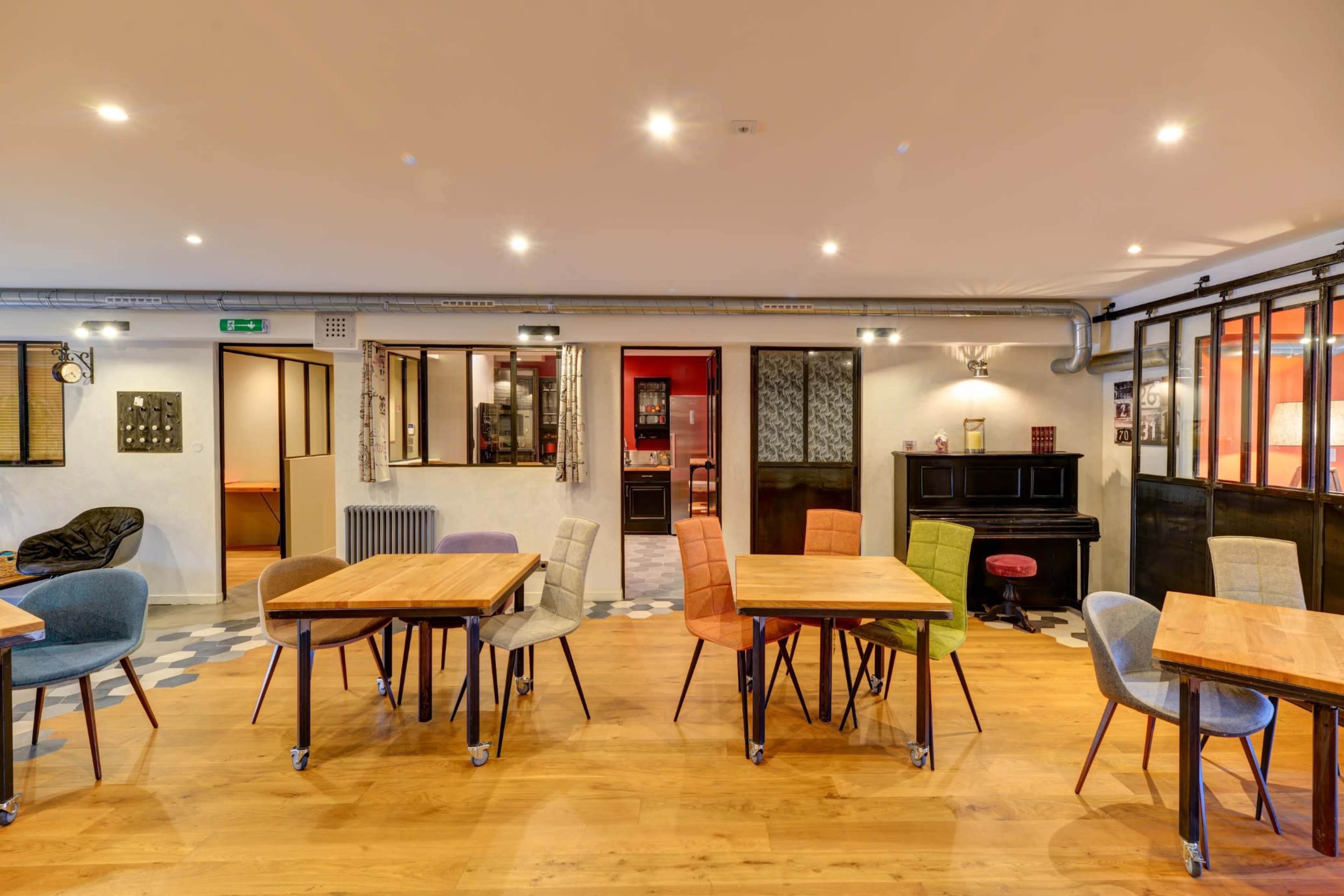 A modern, spacious café interior with multiple wooden tables and colorful chairs arranged on a wooden floor, featuring large windows and soft lighting.