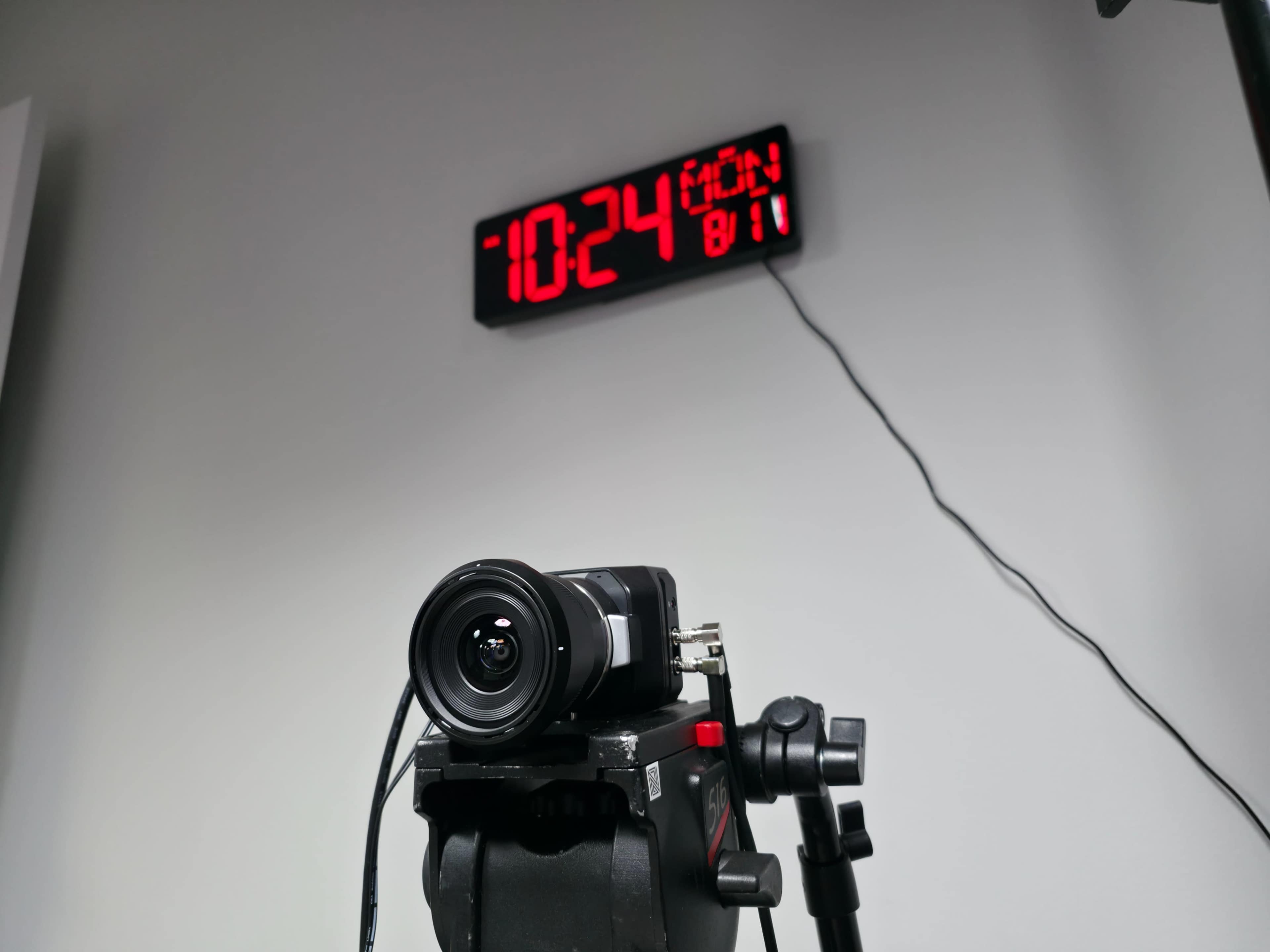 A camera is mounted on a tripod in front of a digital clock displaying the time as 10:24.
