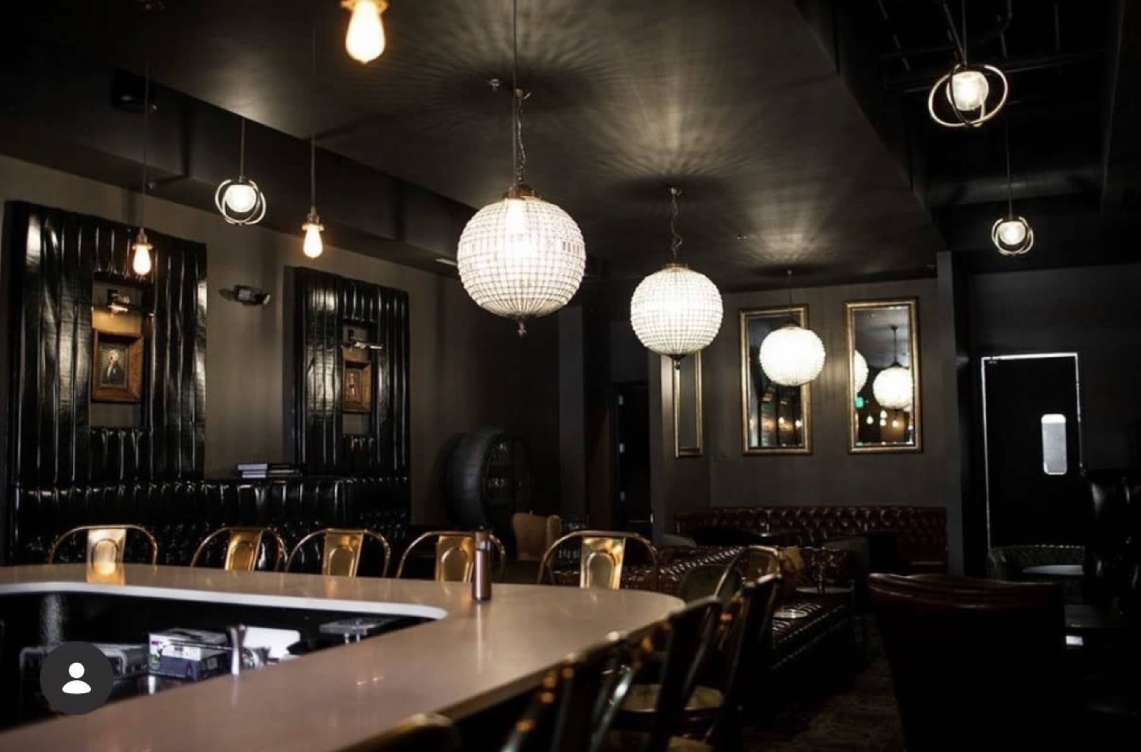 The image shows a dimly lit bar area featuring modern pendant lights, a white countertop, and leather furniture.