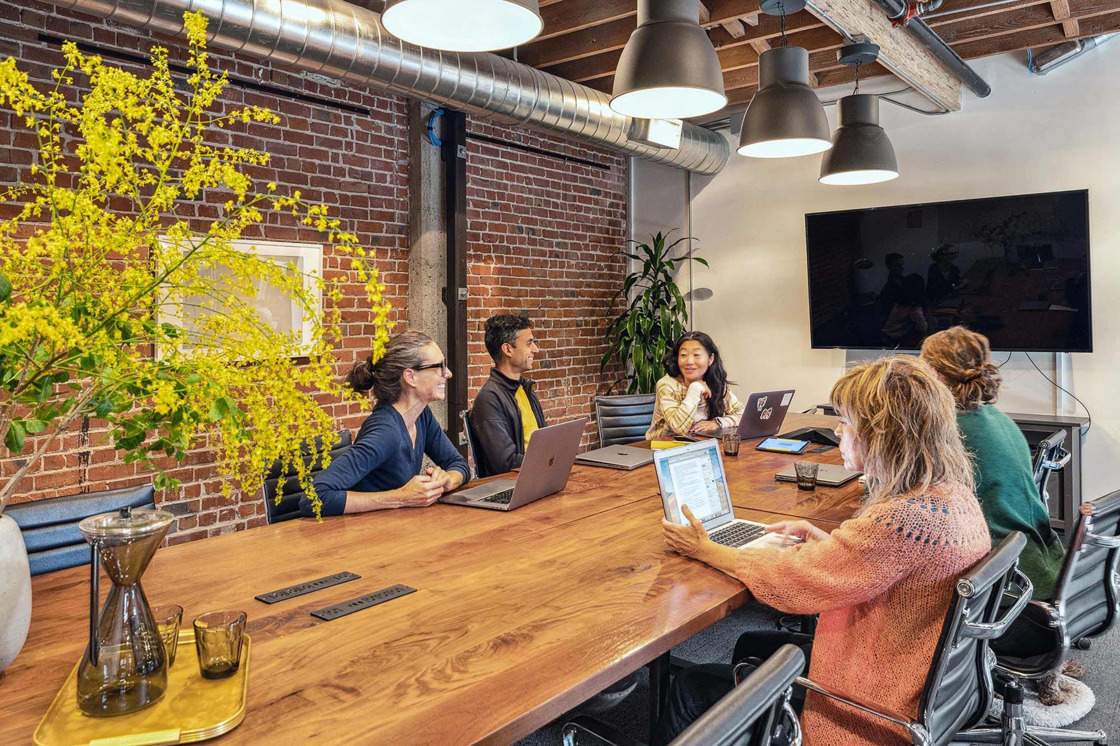 A group of four people is seated around a wooden conference table, discussing while using laptops in a modern meeting room with brick walls and large windows.