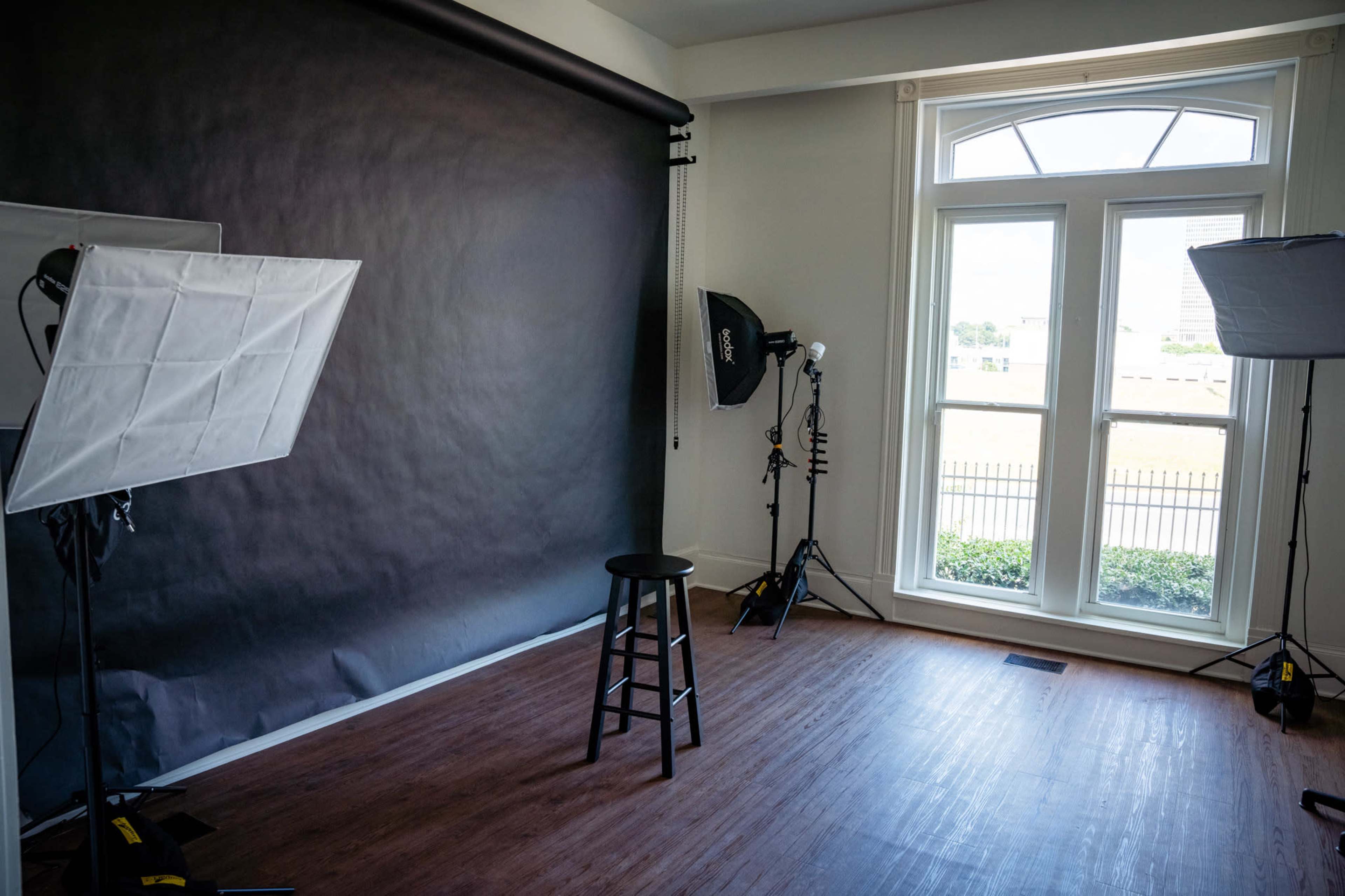 The image shows a photography studio featuring a black backdrop, two light sources, and a stool positioned in front of large windows.