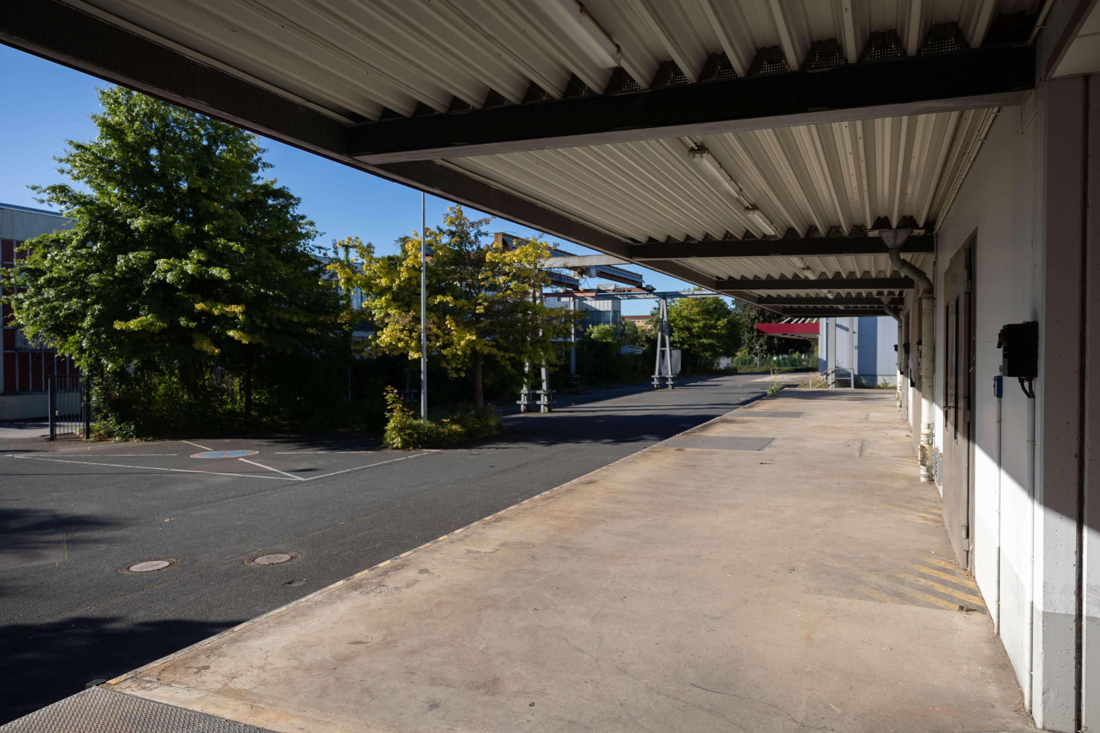 The image shows a shadowed loading dock area with an empty paved space and trees lining the background under clear blue skies.