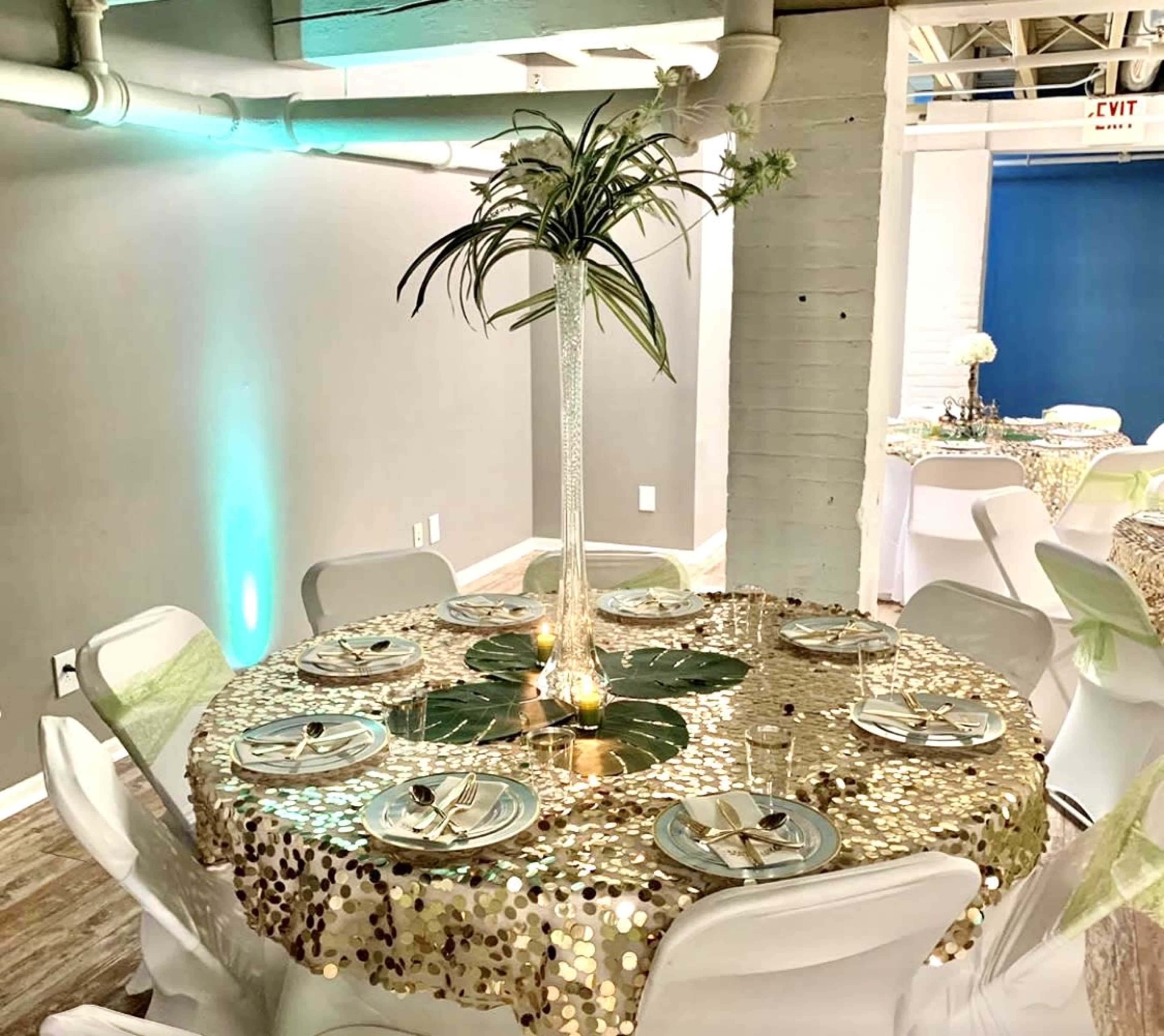 A round table with a sparkling gold sequin tablecloth is set for a formal dinner, featuring white plates, clear glasses, and a tall floral centerpiece.