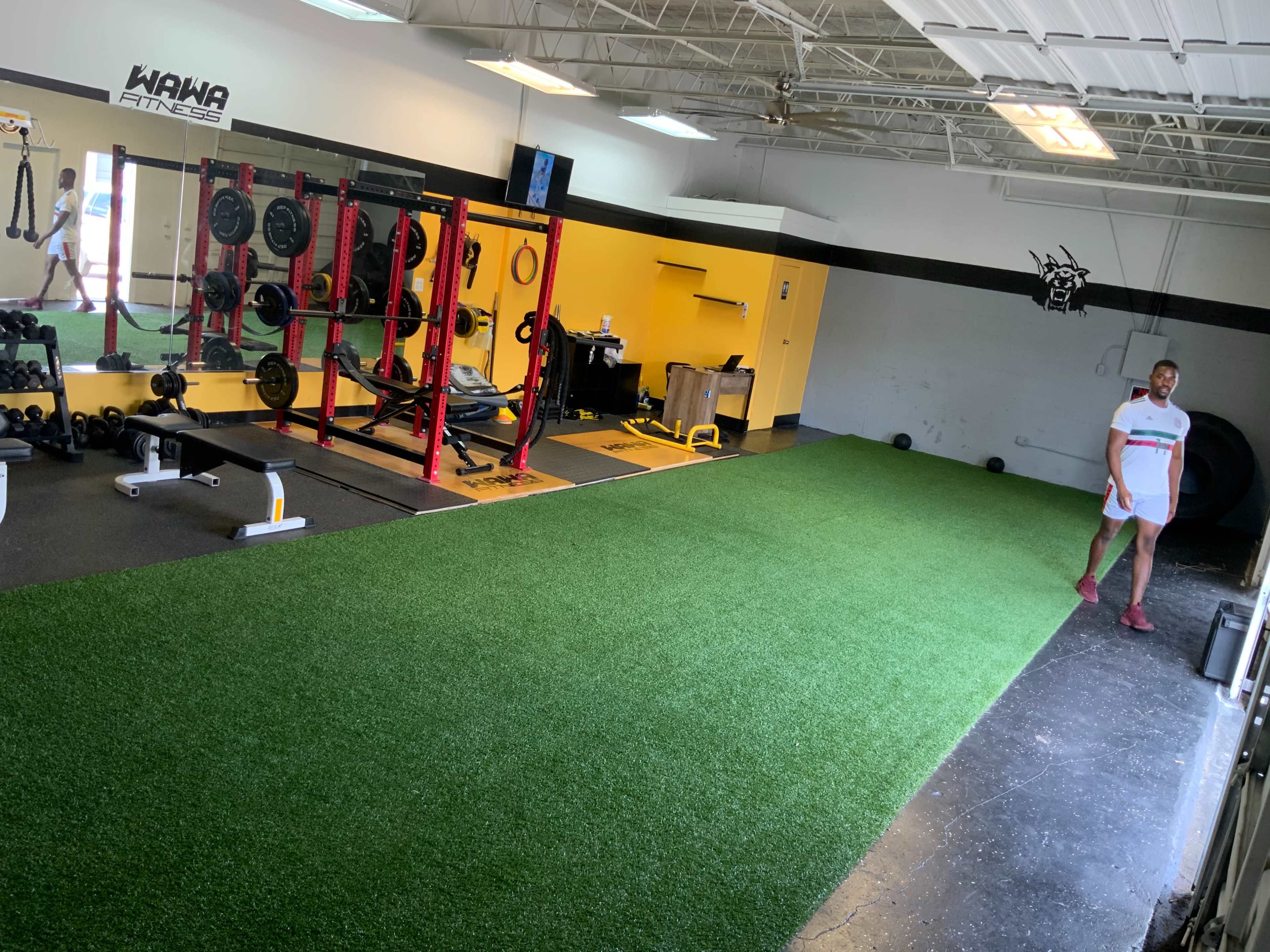 The image shows a gym interior with weightlifting equipment, a section of artificial grass, and a person walking on the gym floor.