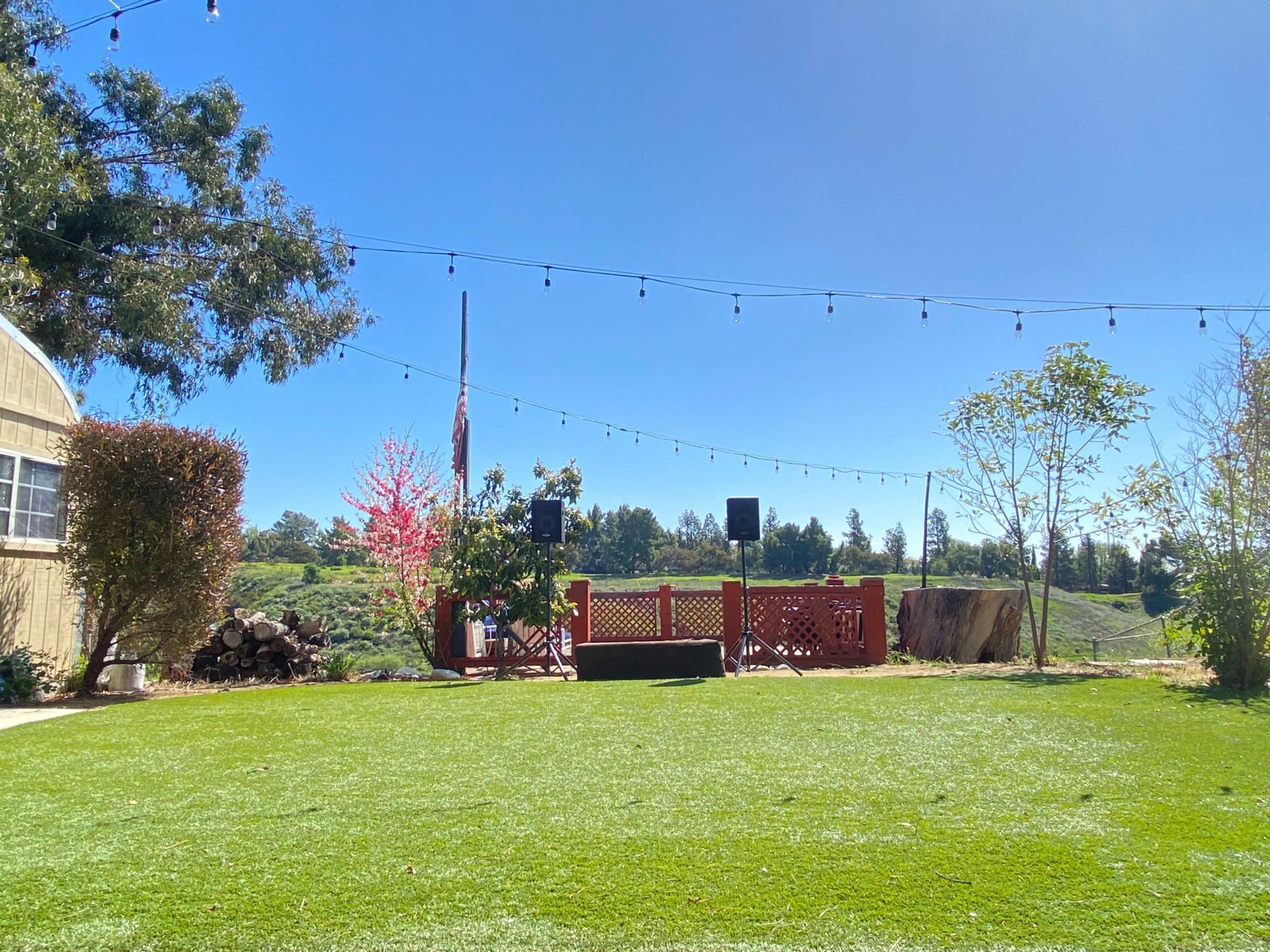 A well-maintained grassy area with a stage and sound equipment set up, surrounded by trees and strings of lights against a clear blue sky.