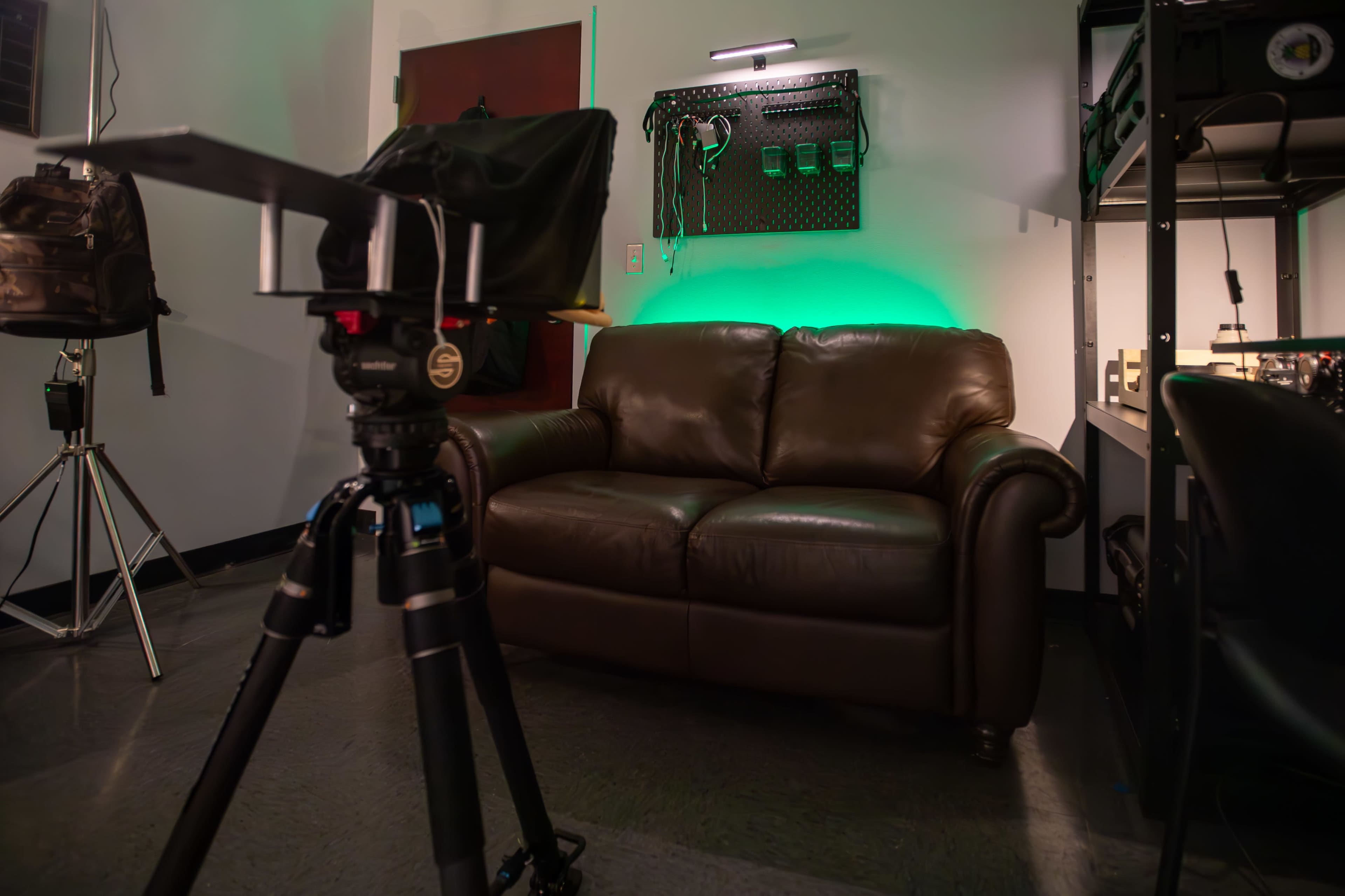 A camera is positioned in front of a brown leather couch in a room with green lighting and various equipment mounted on the wall.