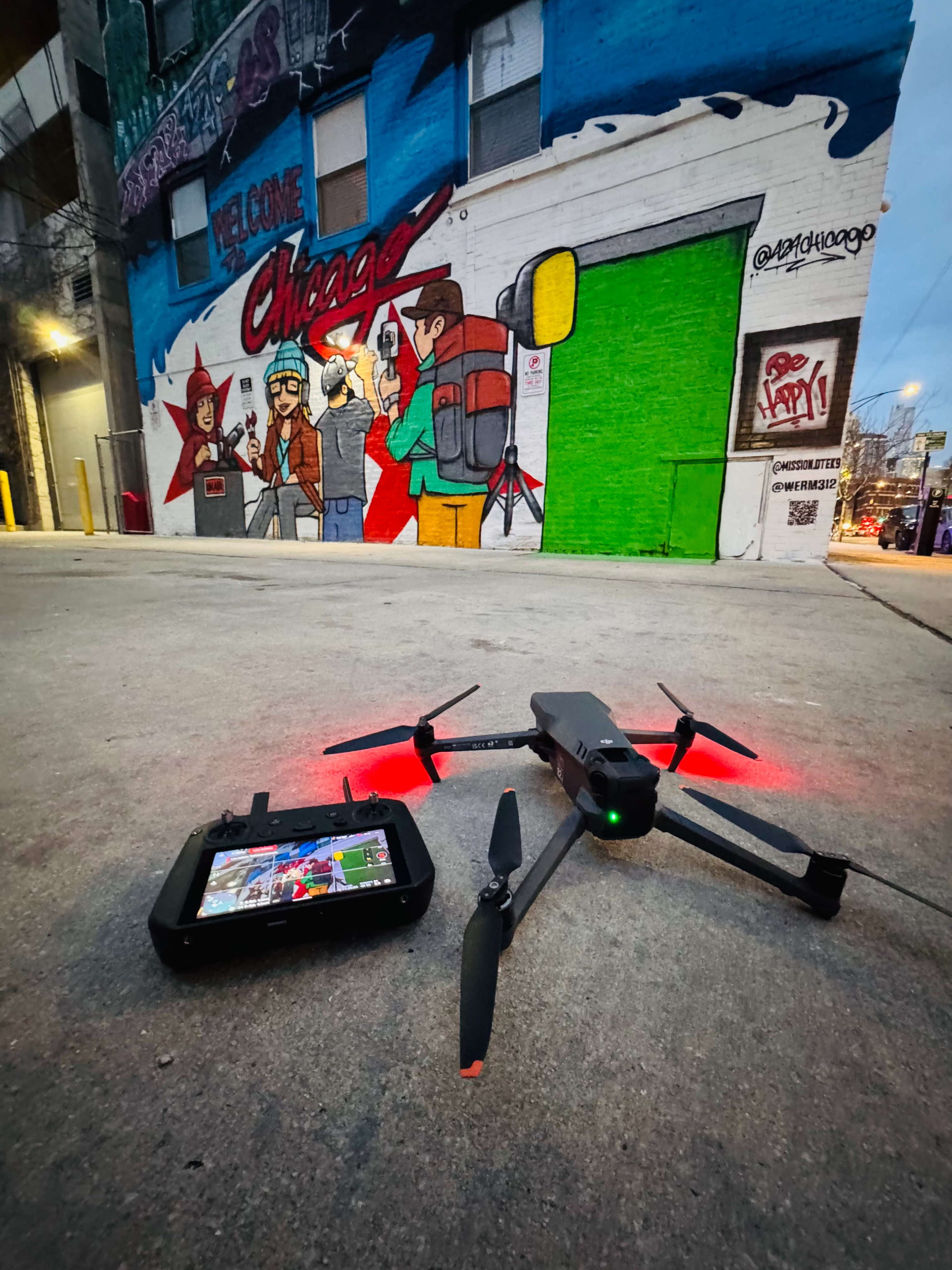 A drone with red lights rests on the pavement in front of a vibrant mural depicting people in an urban setting.