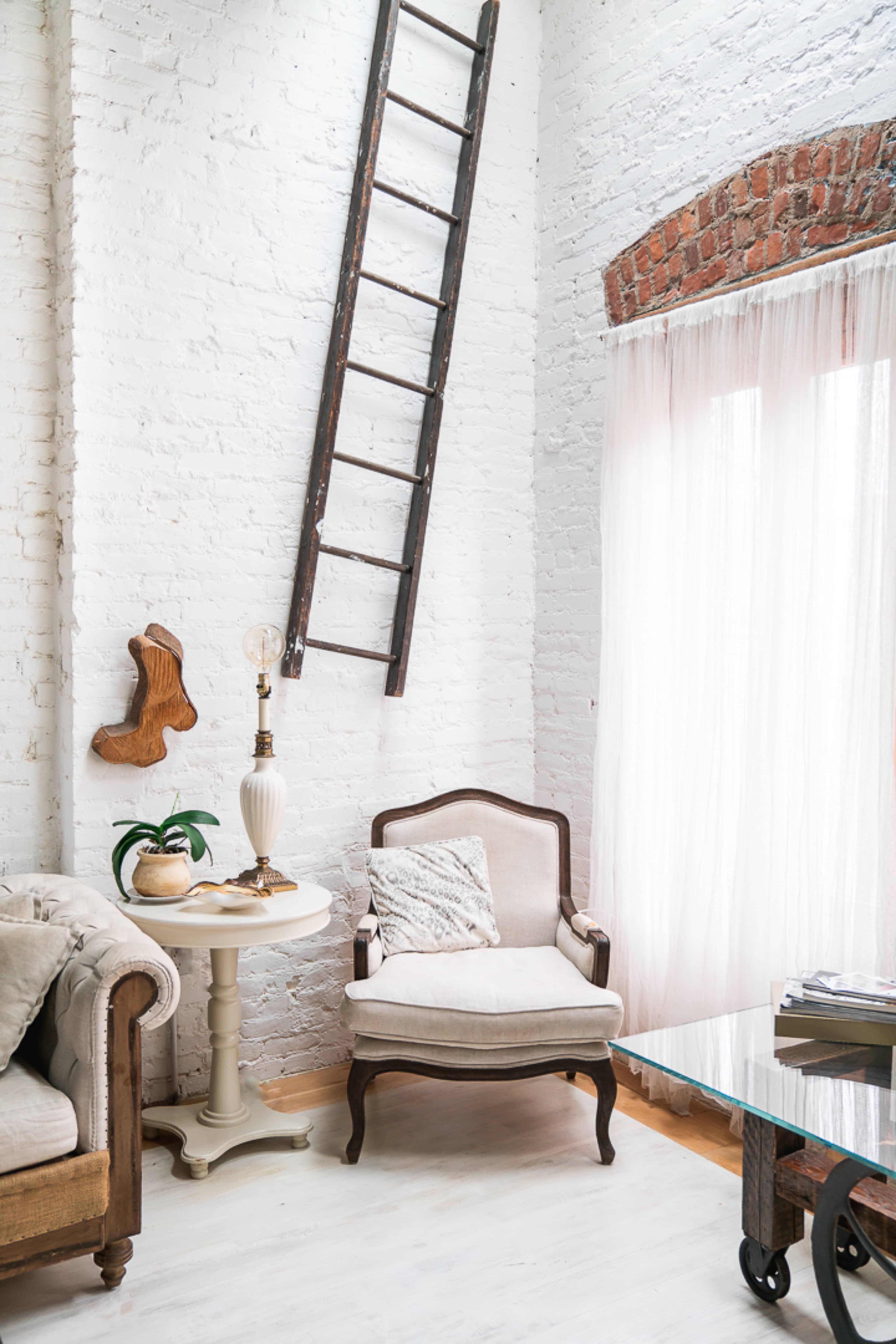 A rustic living room features a wooden ladder leaning against a white brick wall, with a vintage armchair, a side table, and large windows draped with sheer curtains.