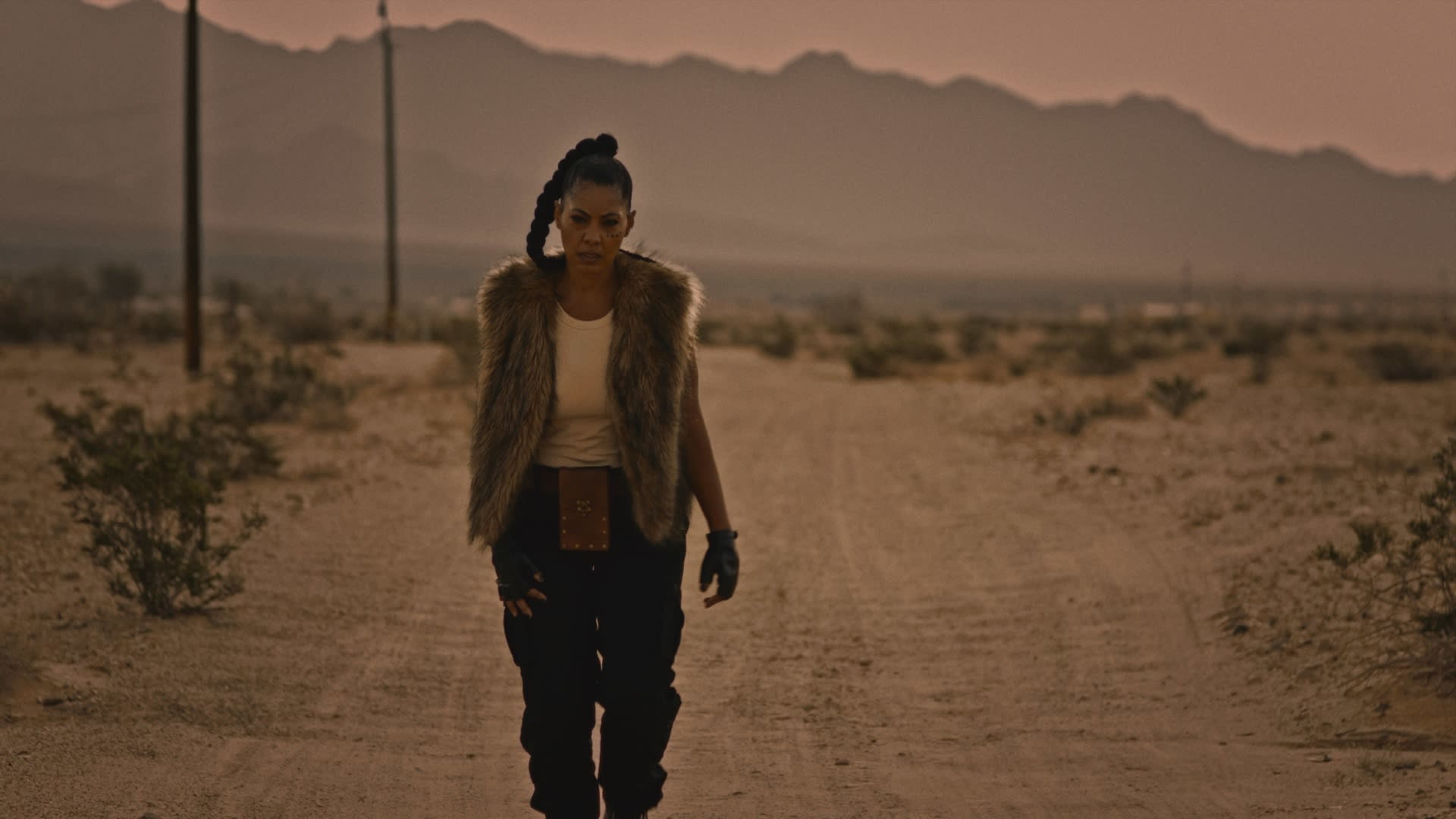 A lone figure walks along a dusty road in a desert landscape with mountains in the background.