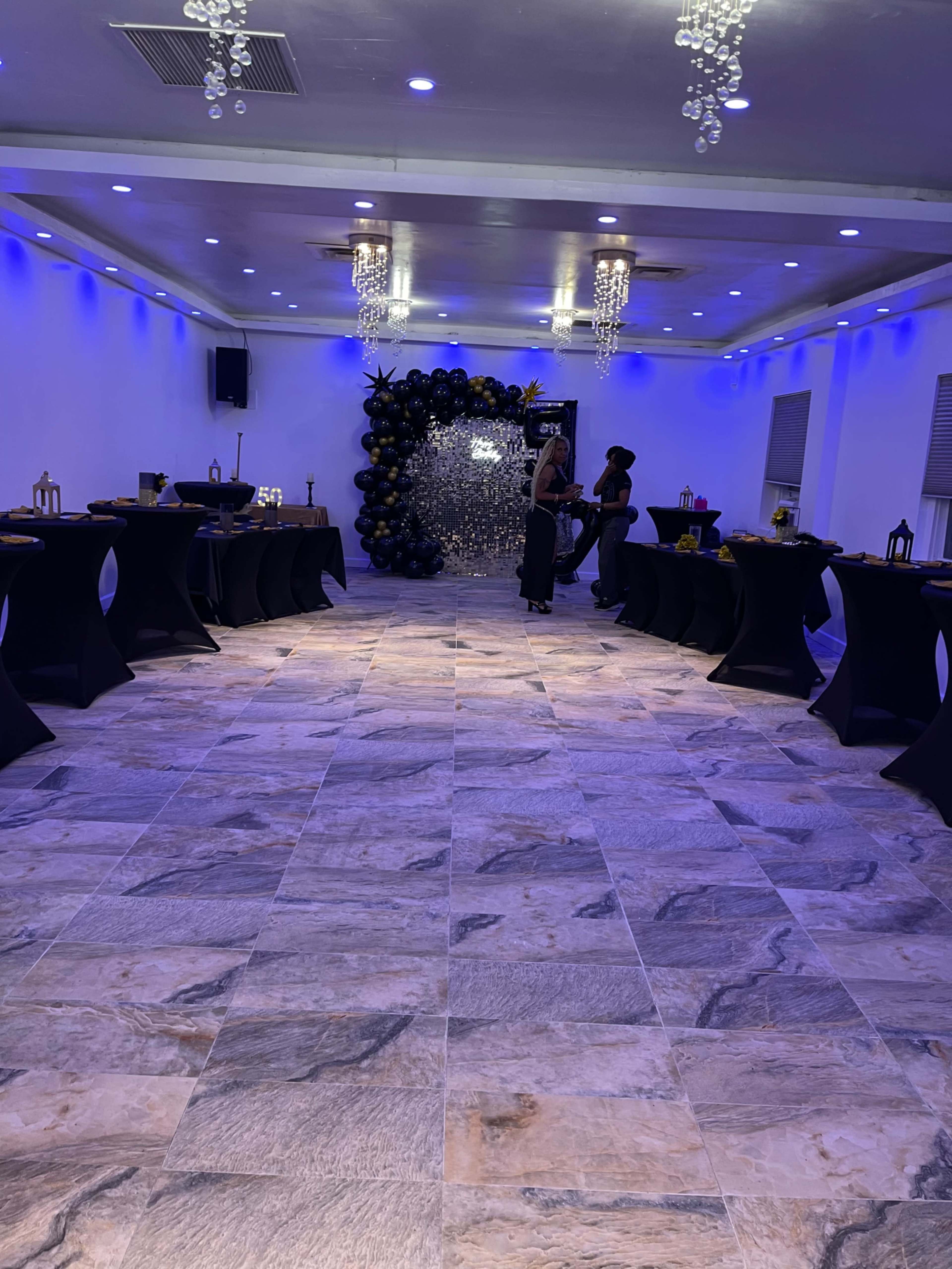 The image shows a spacious event room decorated with black tablecloths, dim lighting, and a backdrop of balloons.